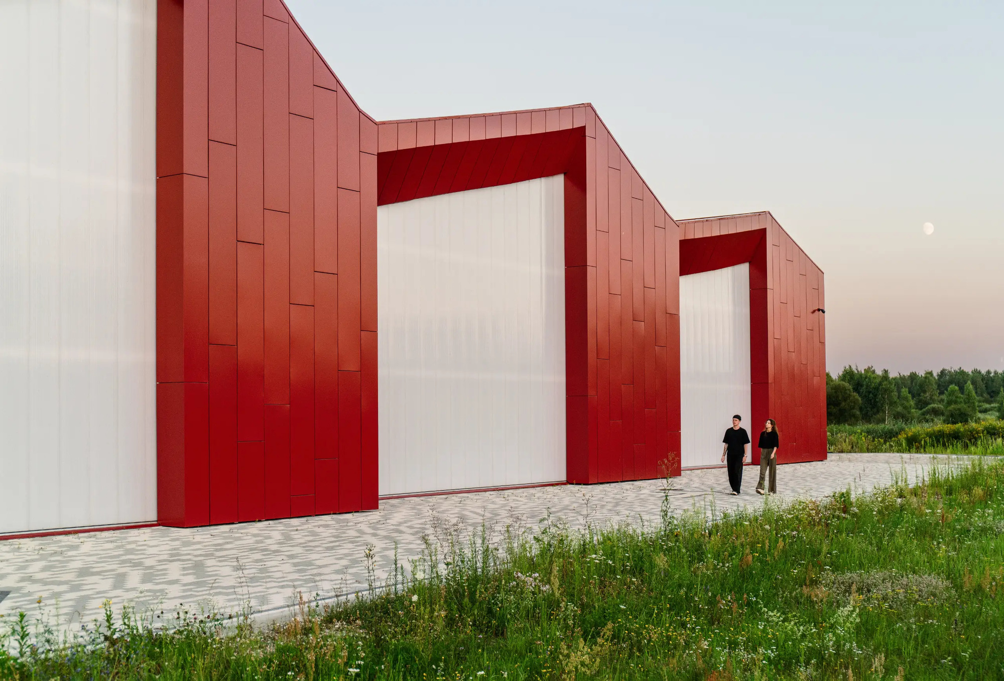 Red metal-clad gabled volumes with recessed white entries and two people walking at dusk