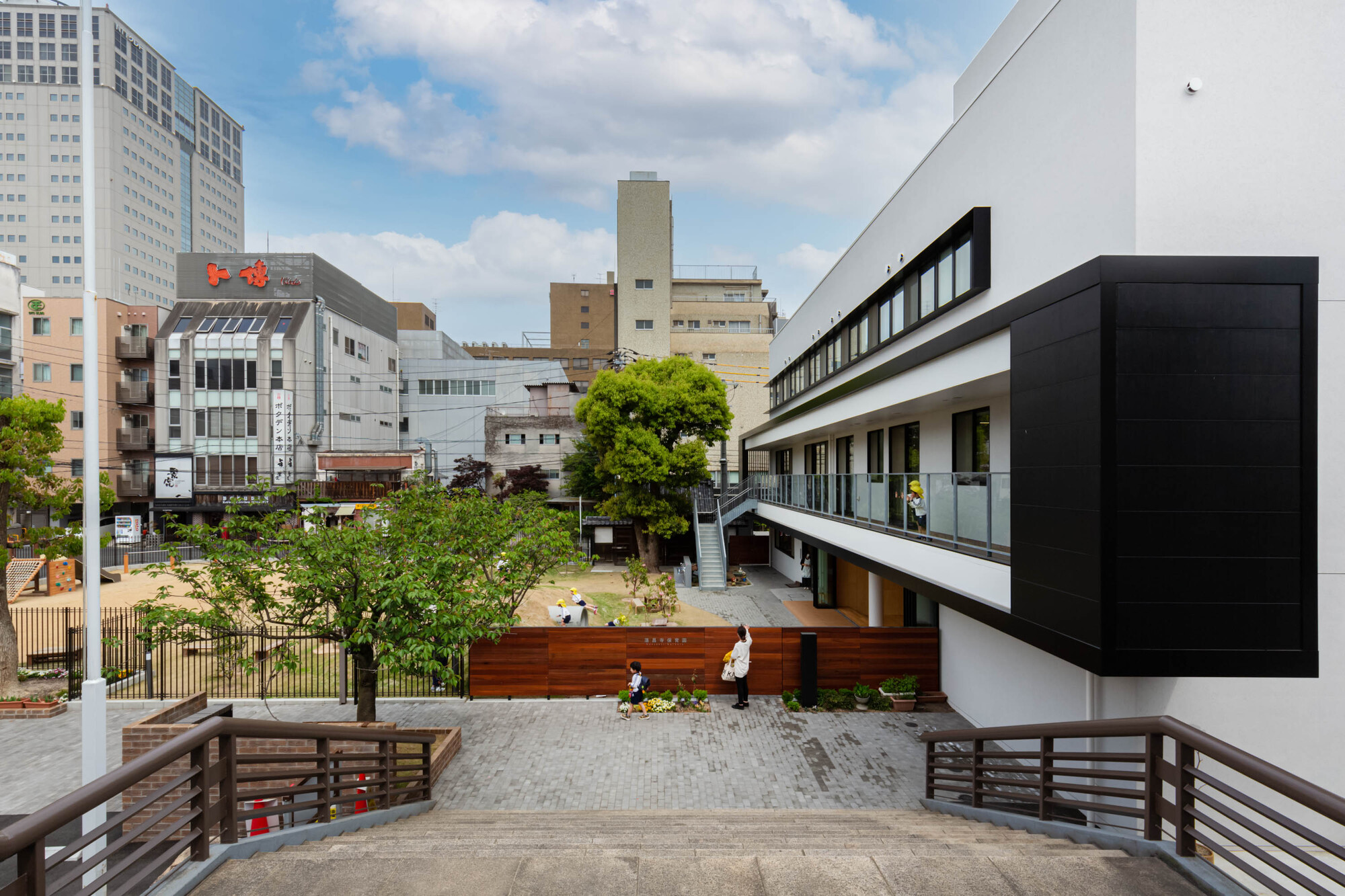 Courtyard view with timber-clad volumes, an elevated walkway and children playing among planted greenery