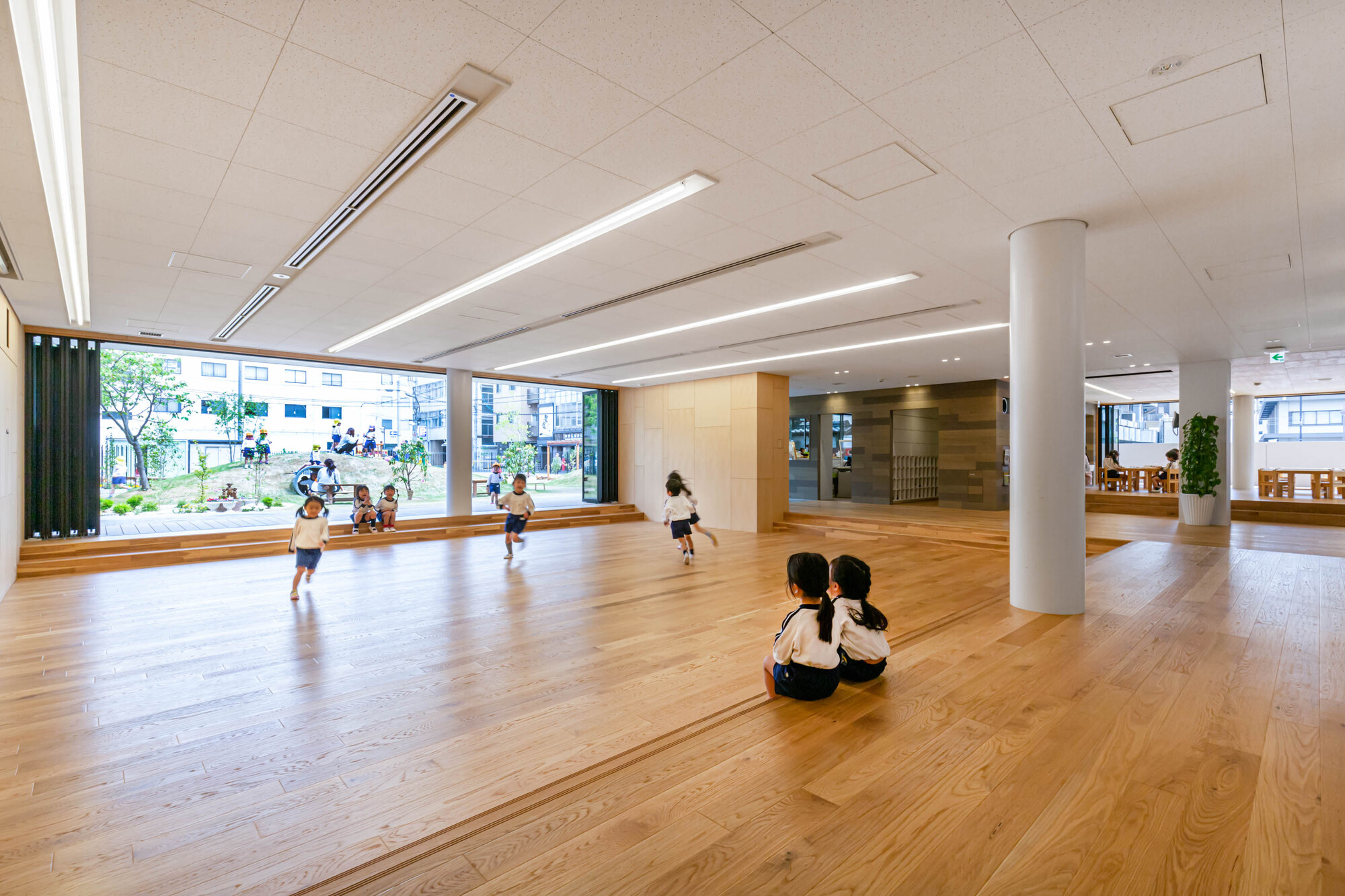 Open timber-floored hall with white cylindrical column and children playing near full-height glazing