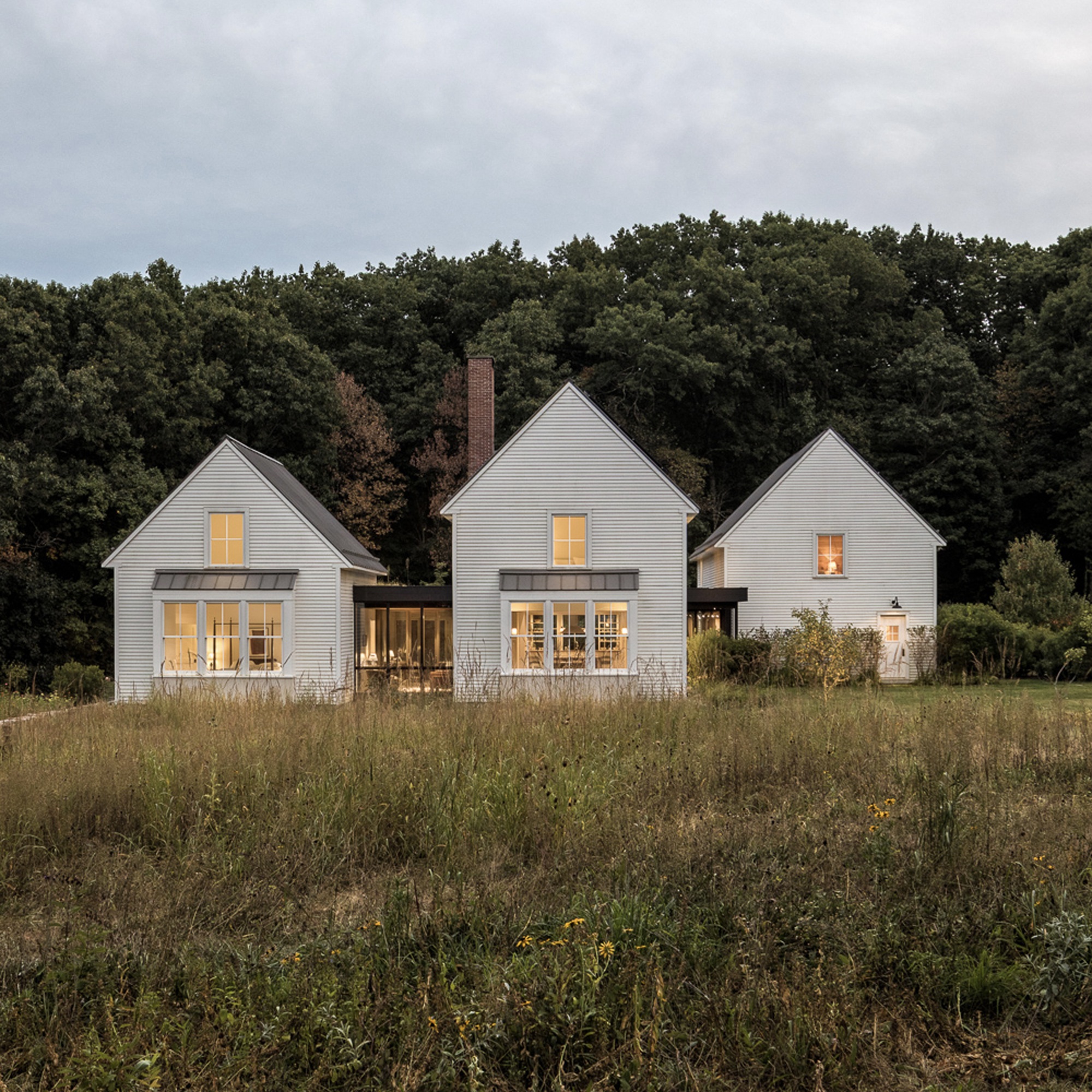 Three white-clad gabled volumes with lit windows across a meadow at dusk