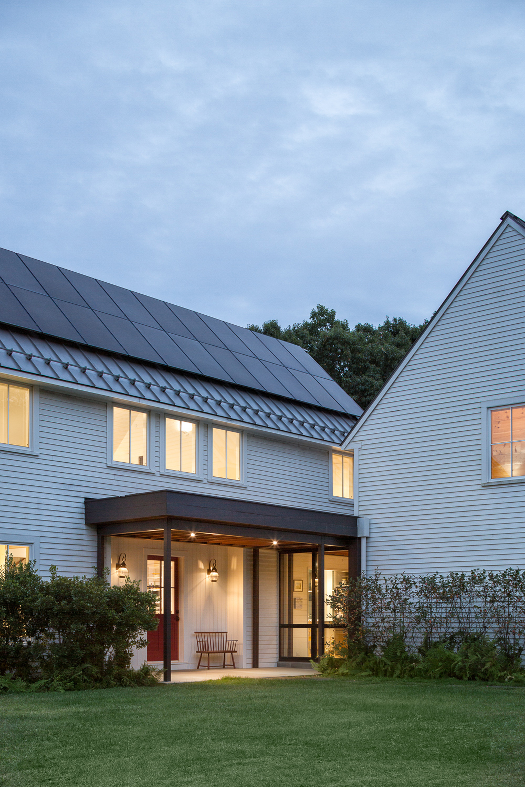Covered porch with timber posts beneath white siding and solar panels on the gabled roof at dusk