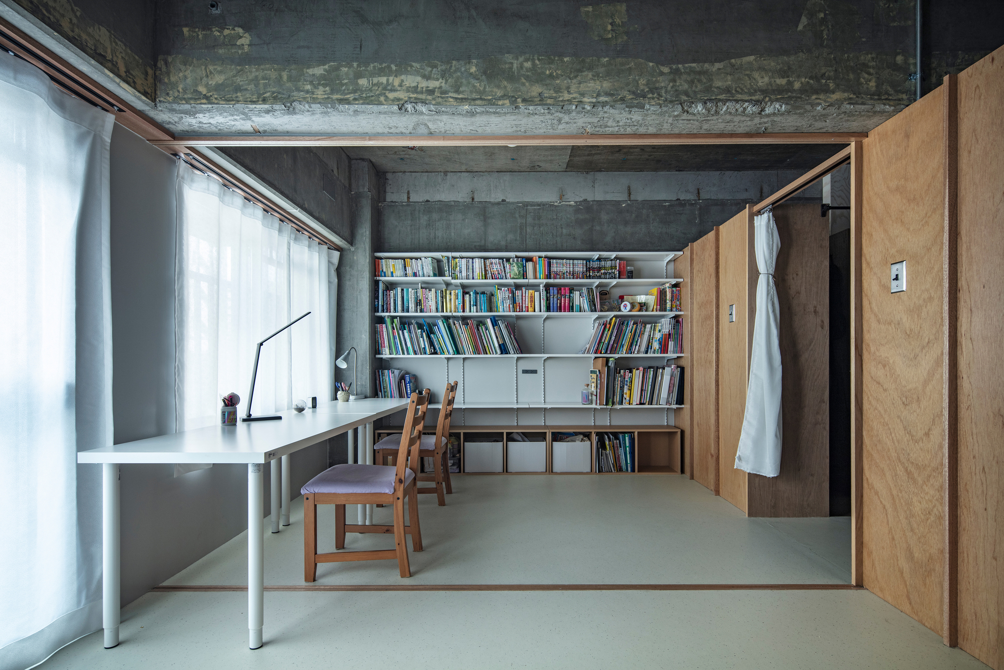 Work area with white desk and open bookshelf against raw concrete walls under natural light from curtained windows