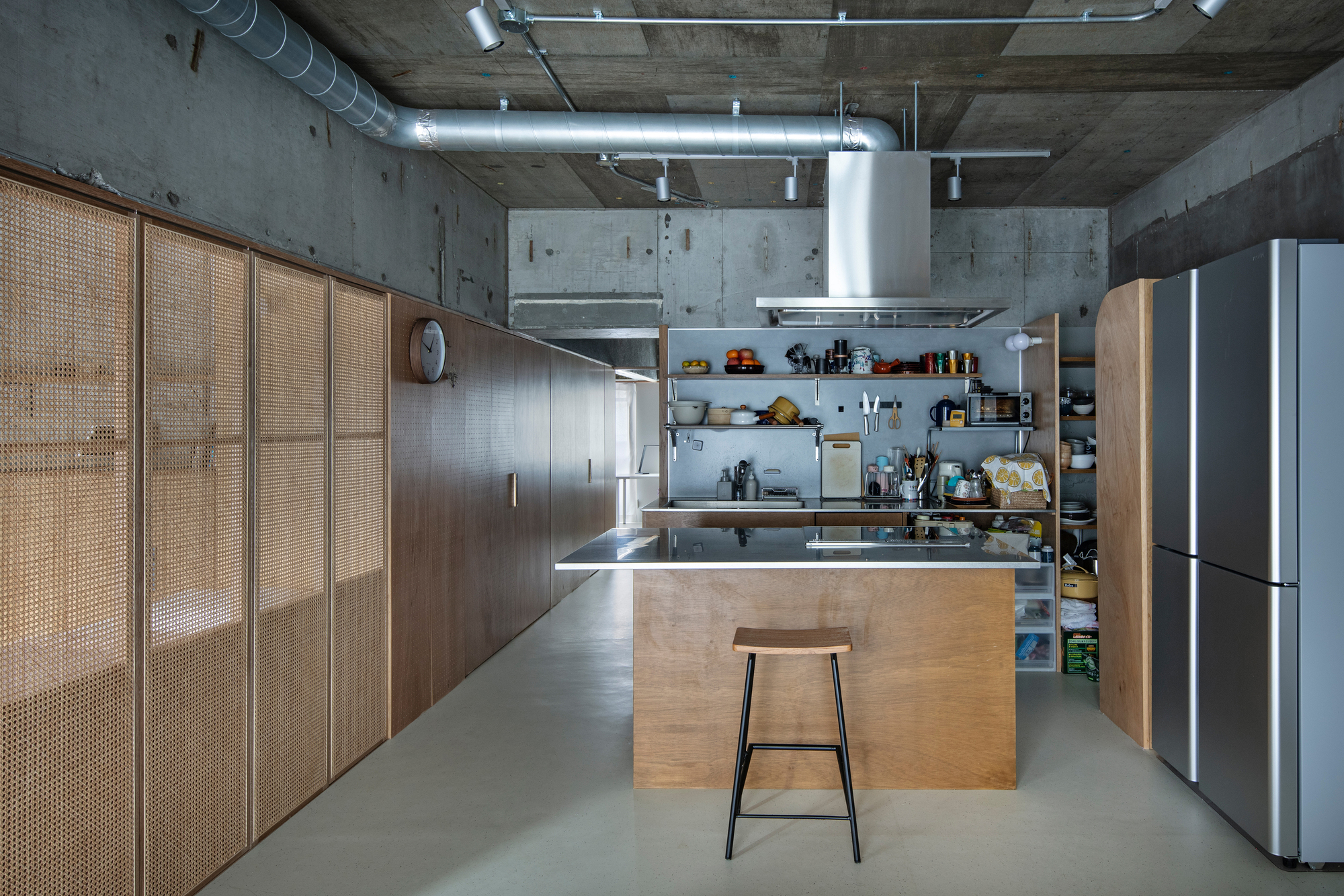 Kitchen island with steel countertop and single stool beneath exposed concrete ceiling with suspended ventilation hood