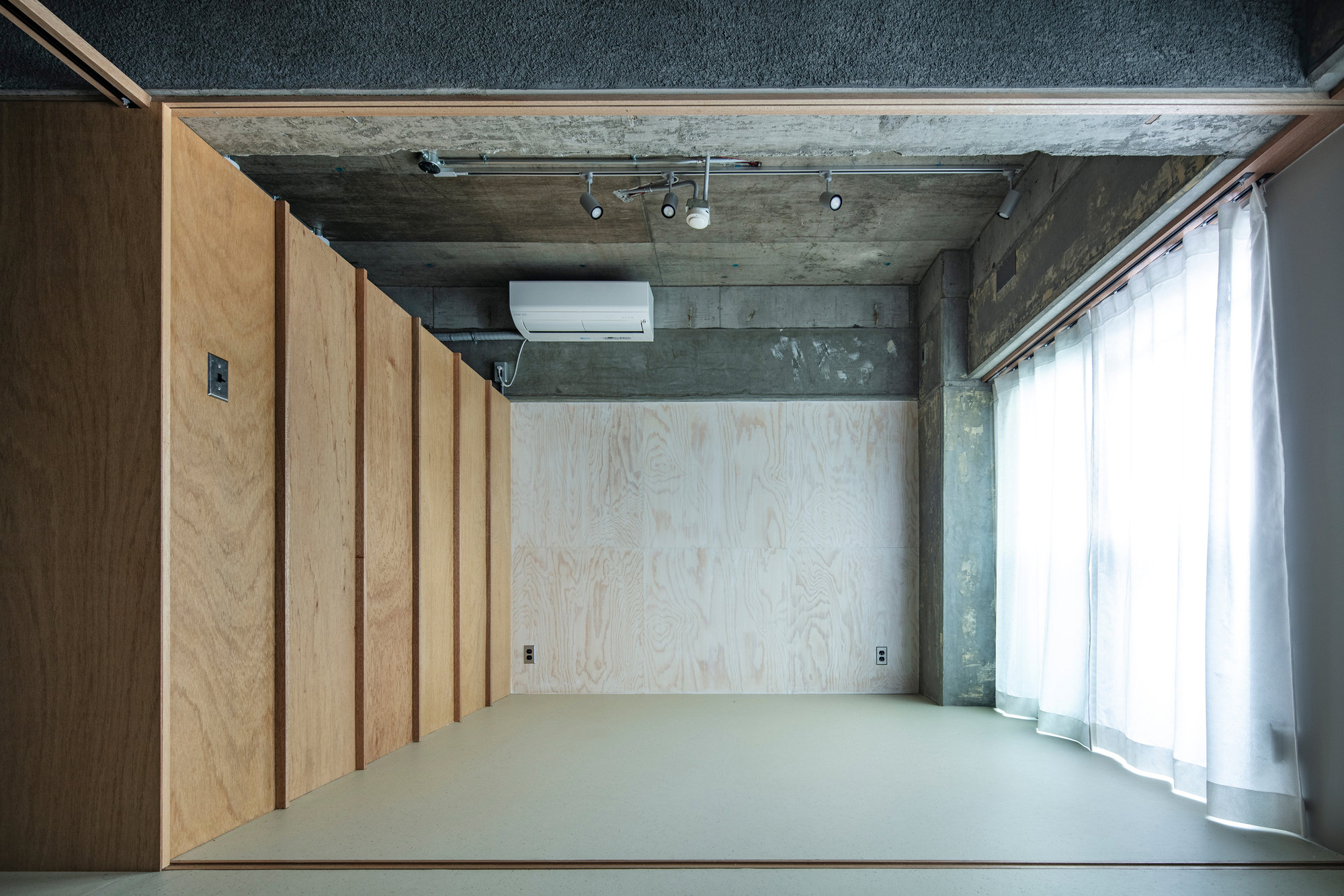 Top-down view of plywood-clad storage wall and grey resin floor lit by diffused daylight through sheer curtains