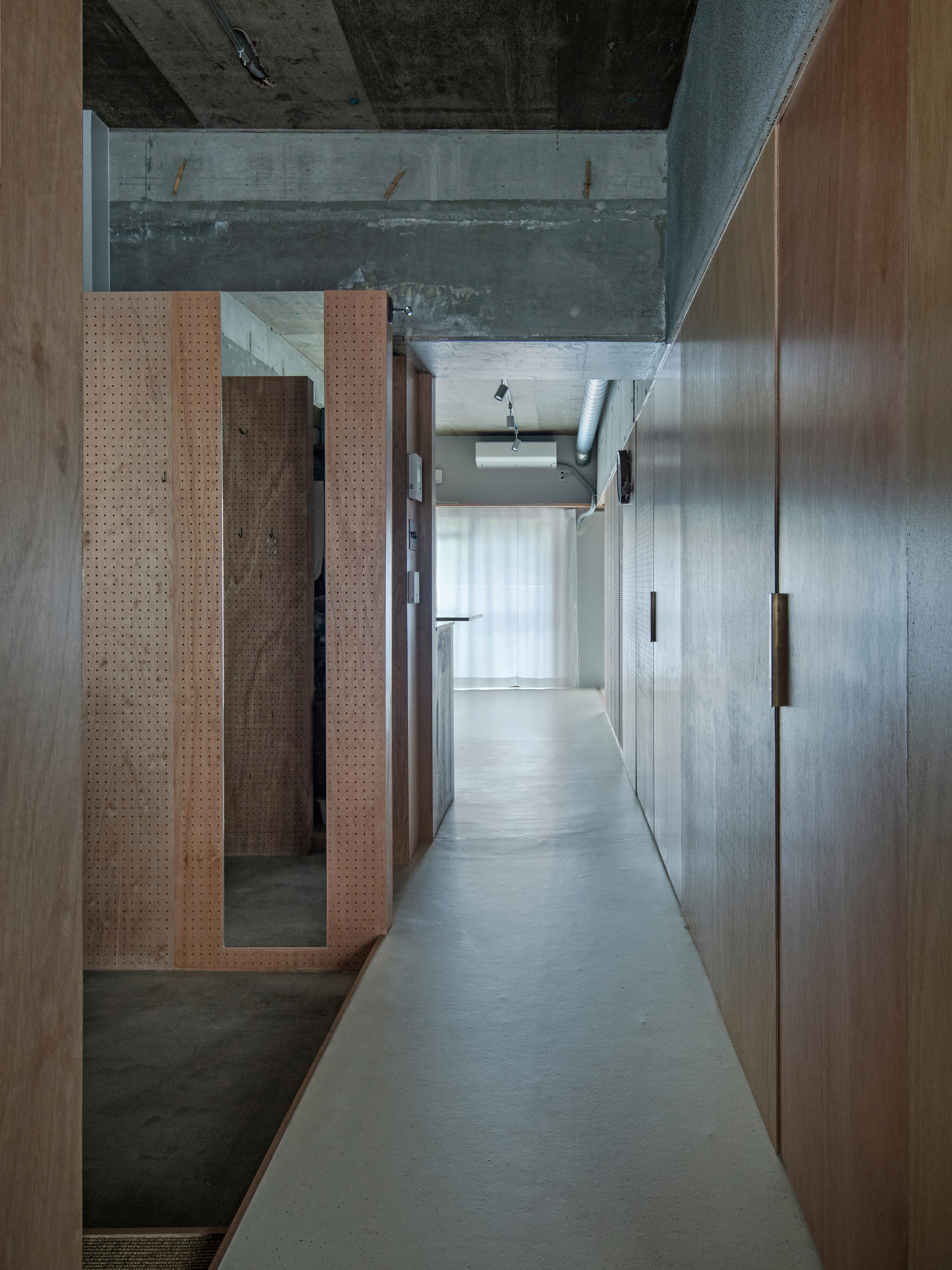 Narrow corridor with perforated timber panel and concrete walls leading toward a backlit curtained opening