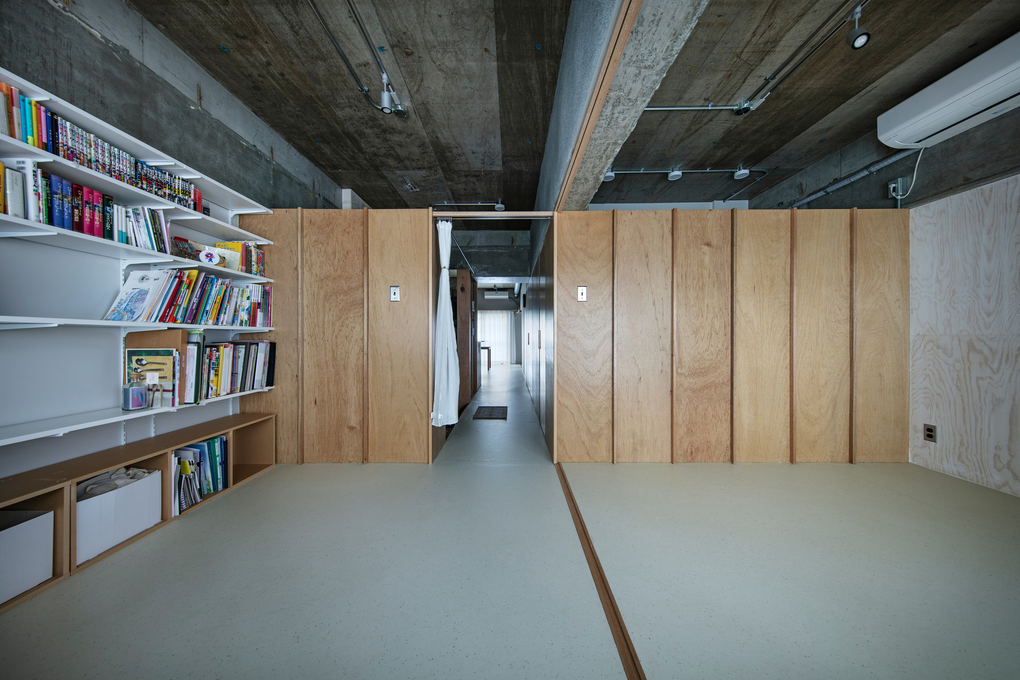 View through wooden partition doorway into hallway with bookshelf and pale flooring under concrete ceiling