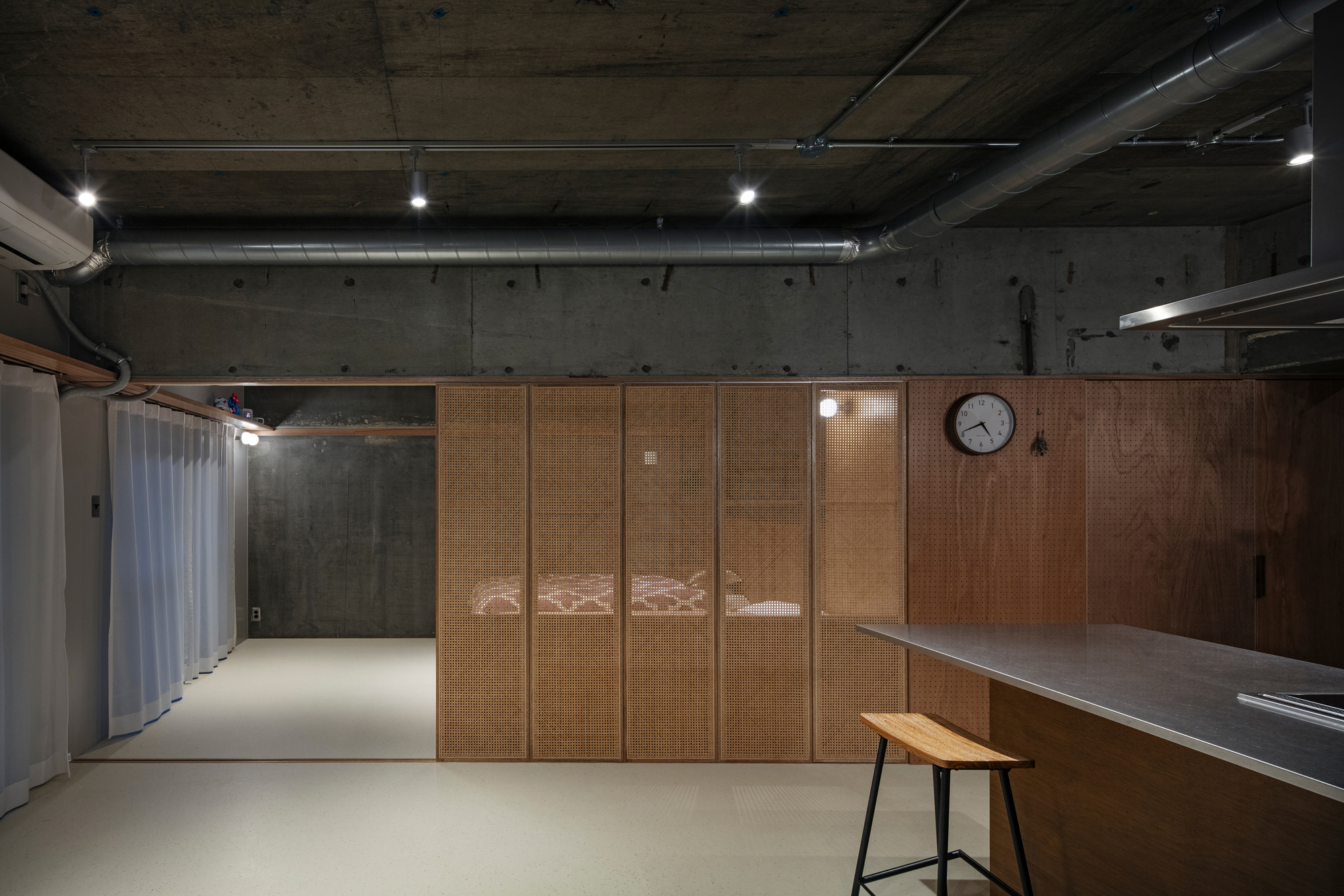 Timber and cane partition wall with round clock, exposed concrete structure and ducting overhead