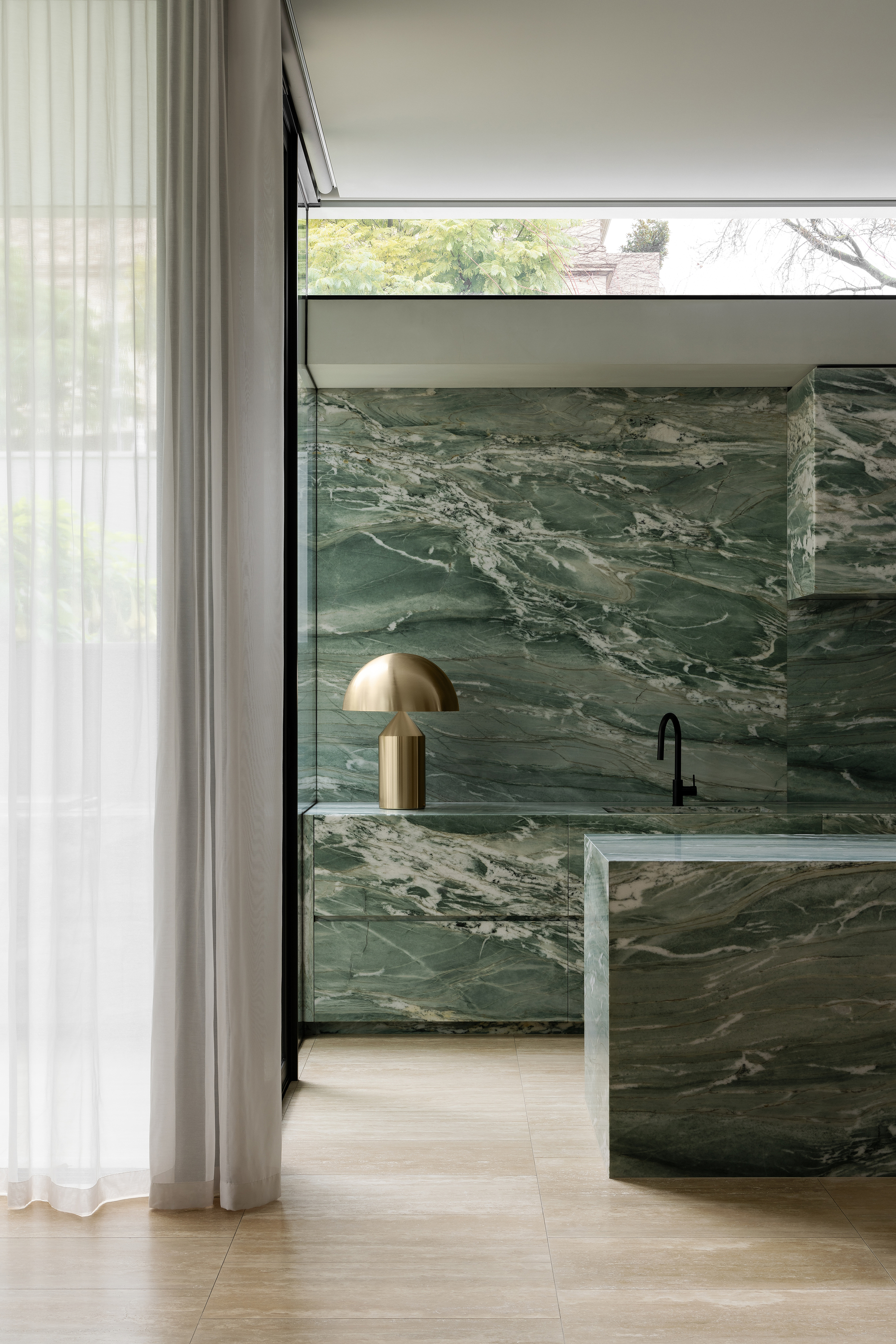 Kitchen island clad in green veined marble with a brass table lamp beside sheer curtains