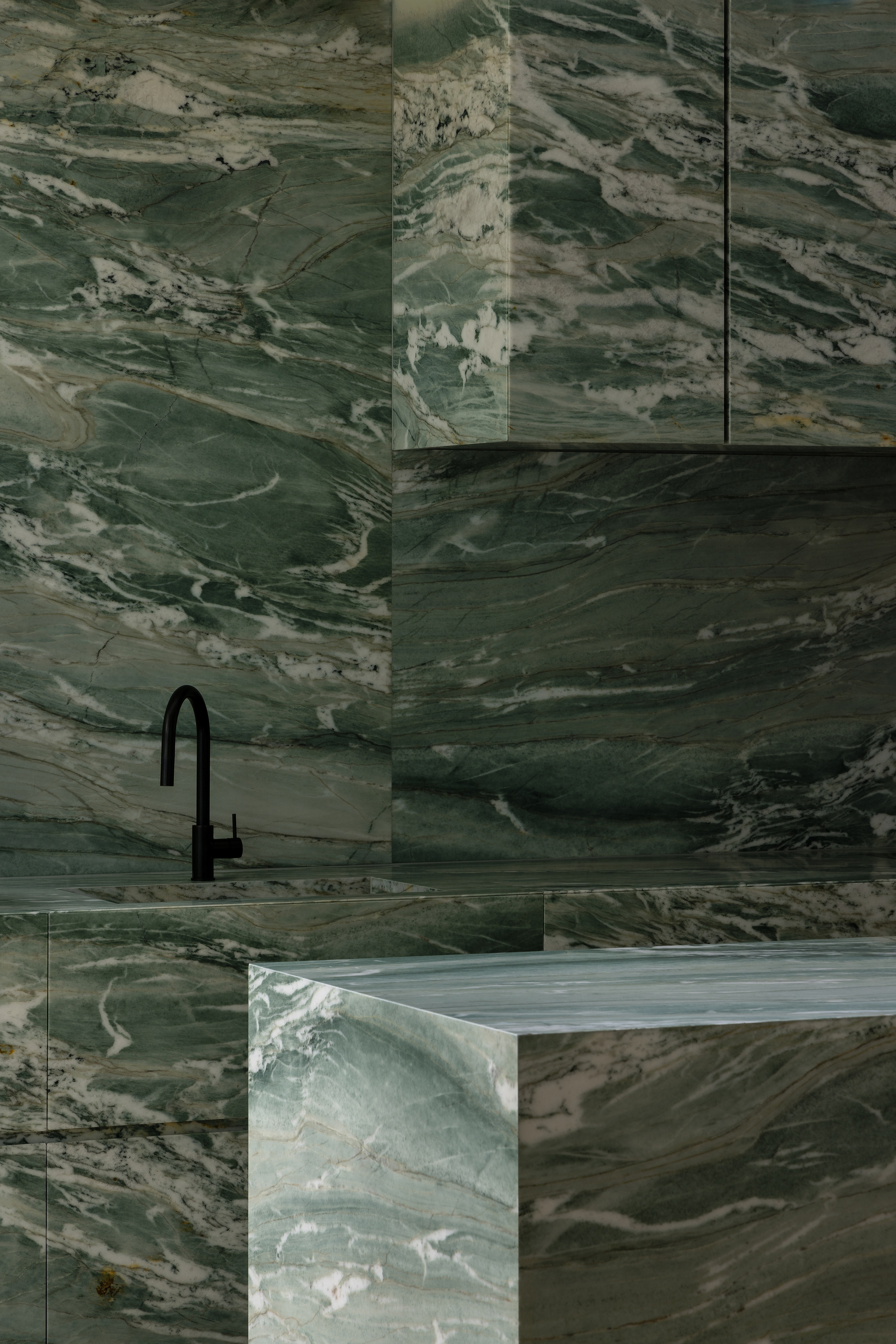 Corner detail of green veined marble surfaces meeting around a black tap fixture and rangehood