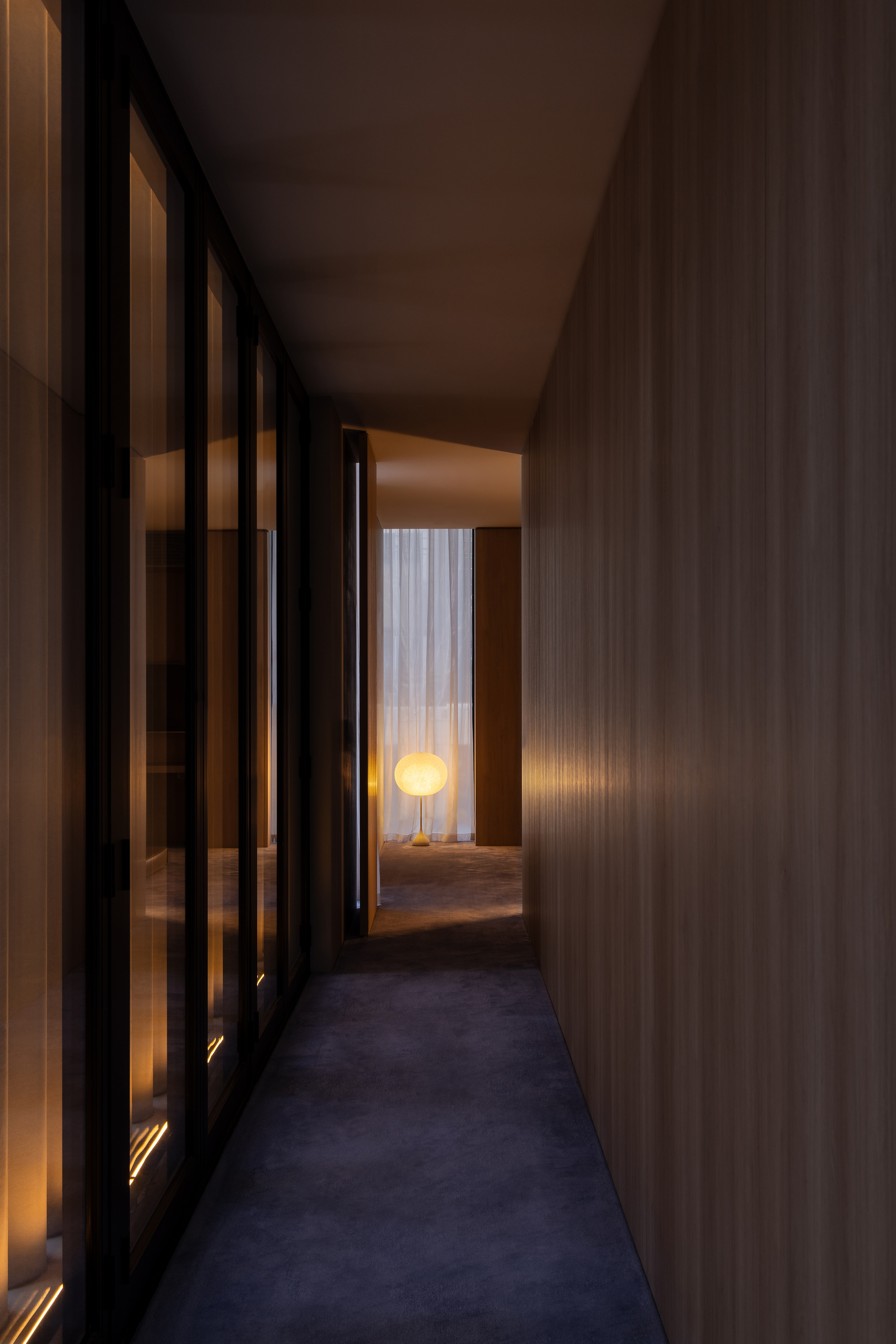 Timber-clad corridor with glass doors leading to a room with illuminated floor lamp