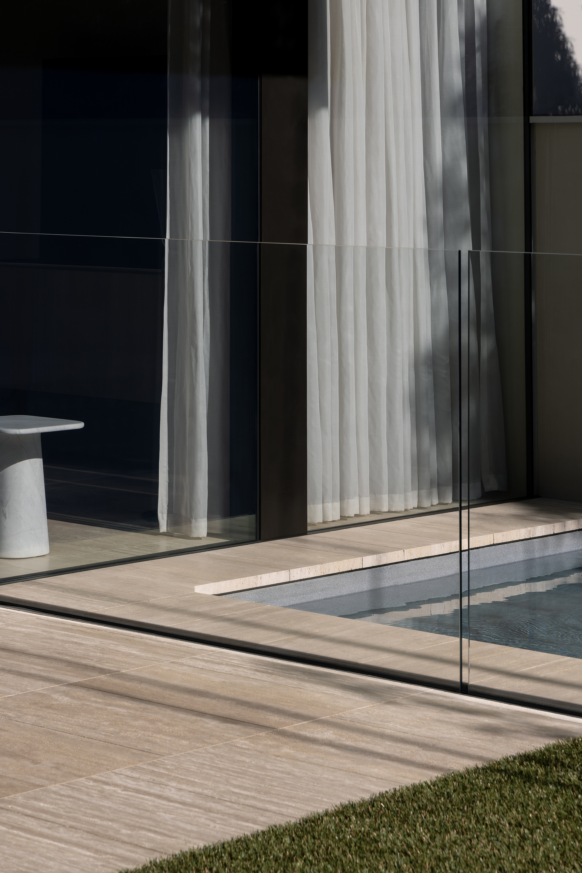 Glass balustrade and travertine deck edge beside a pool with sheer curtains beyond