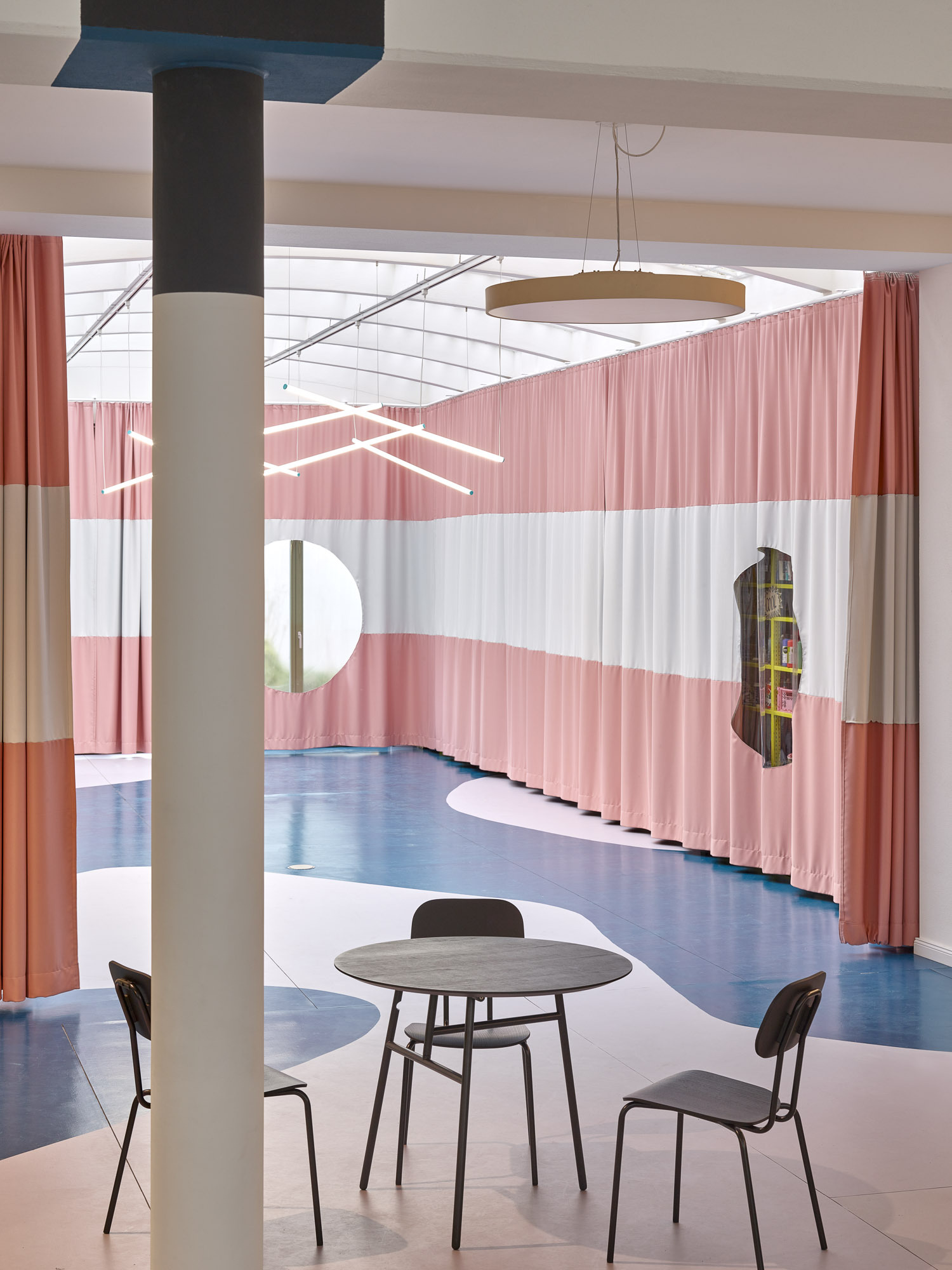 Interior with striped pink curtain partitions and round table beneath a coffered skylight and suspended ring light