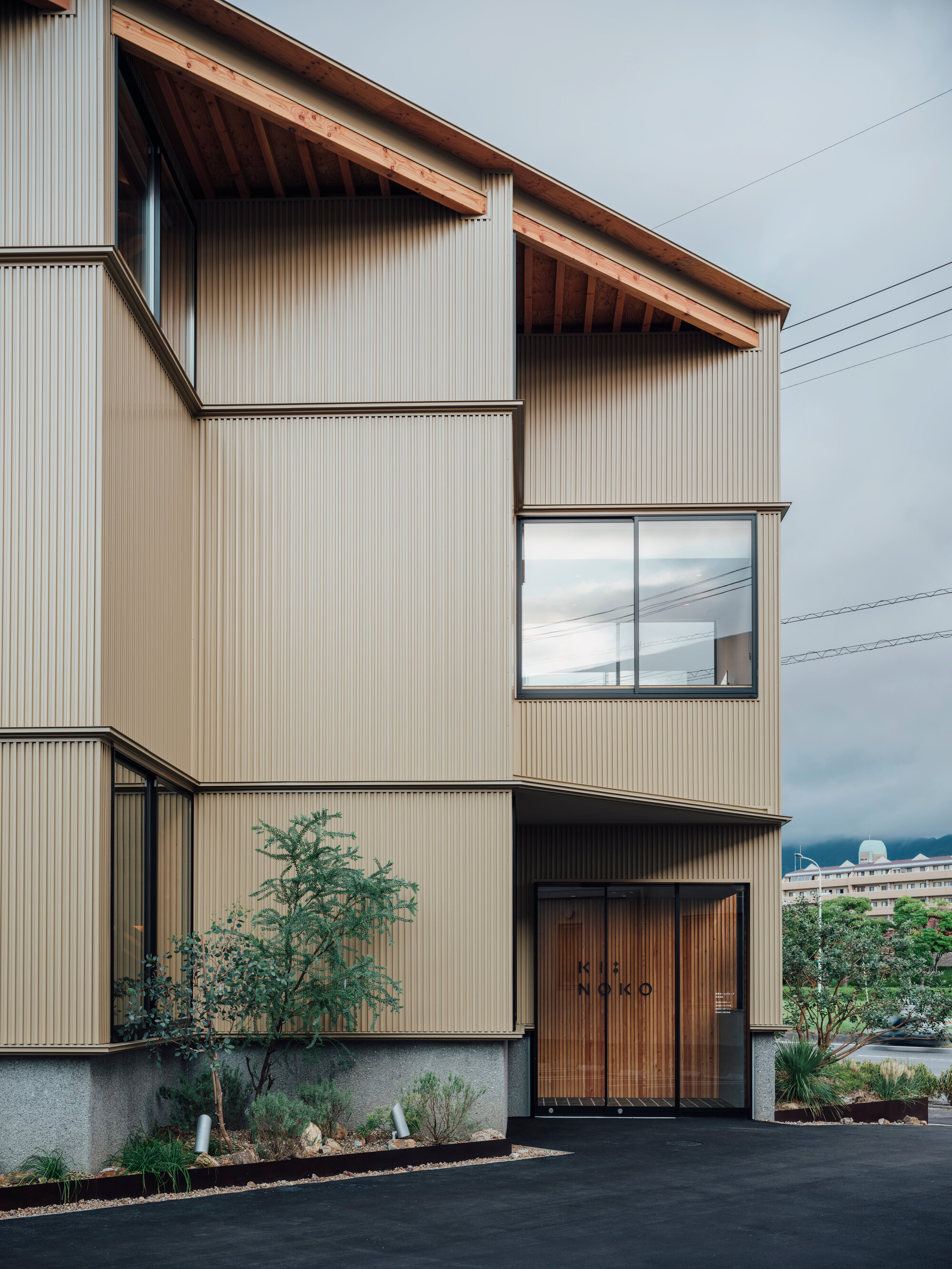 Front elevation showing corrugated metal walls and timber entrance doors with planted foreground under cloudy skies