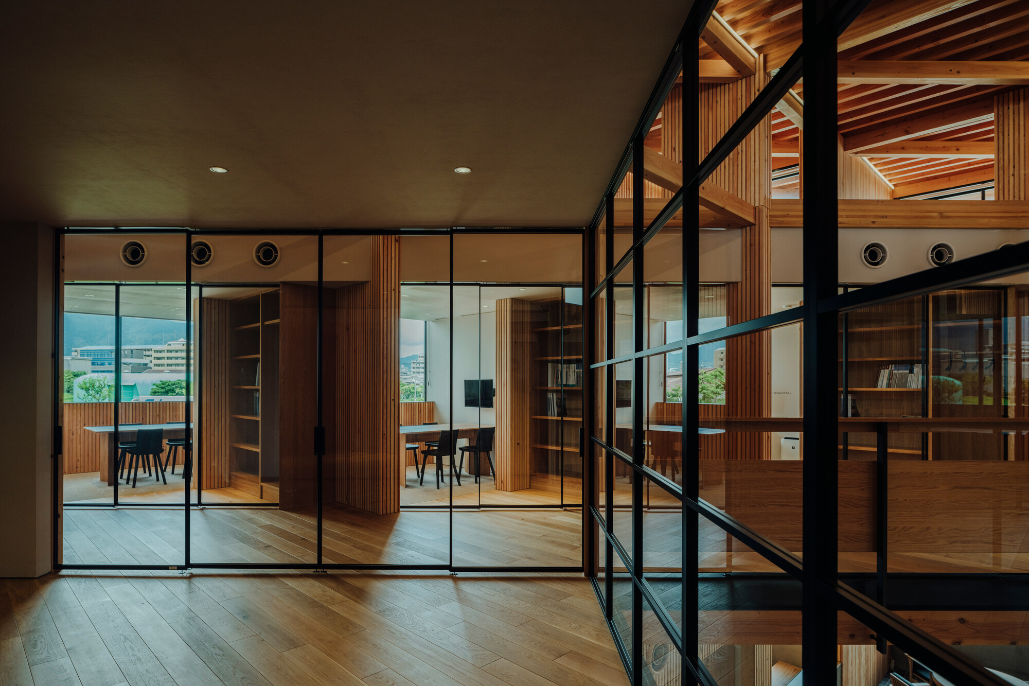 Interior hallway with black-framed glass partitions revealing adjacent timber-clad rooms and exposed joists in evening light