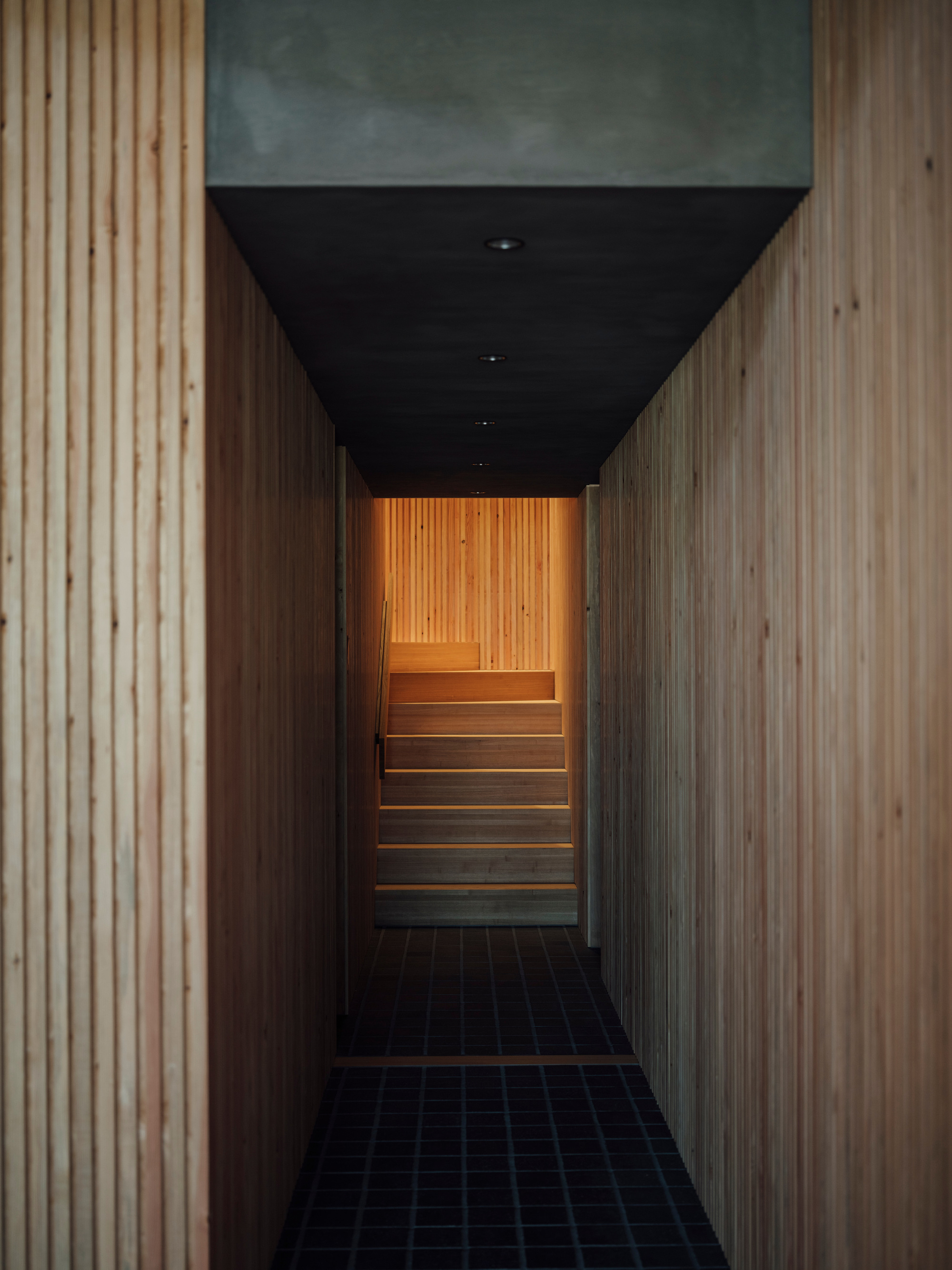 Narrow corridor with black ceiling and blue-tiled floor leading toward illuminated timber staircase at end
