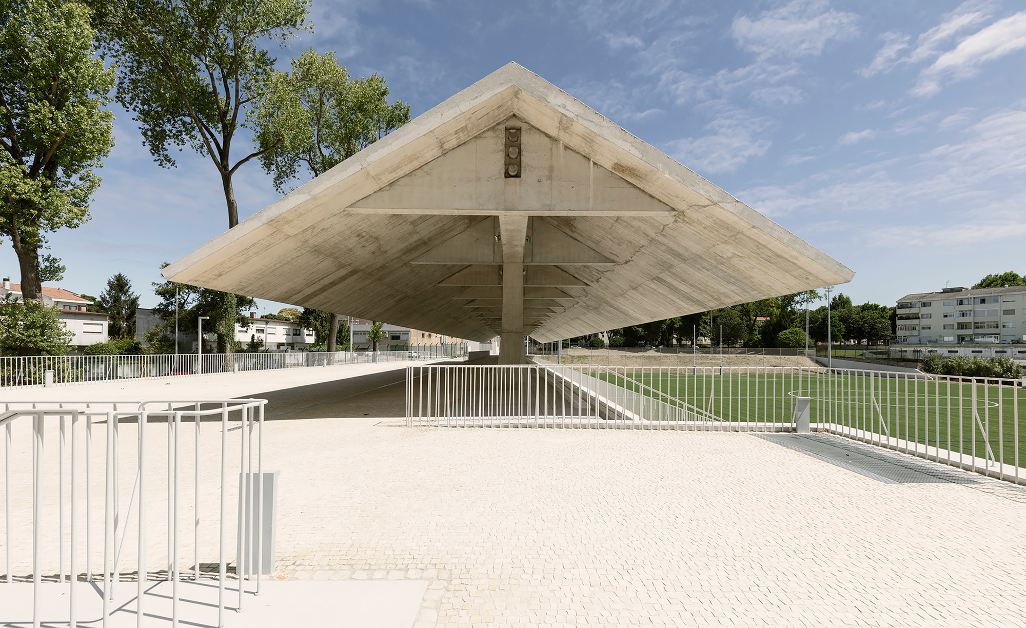 Cantilevered concrete roof with exposed formwork hovering above white railings and a football pitch