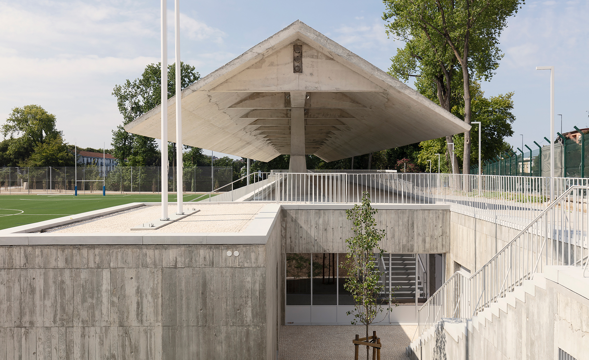 Concrete pavilion entrance set into a bermed podium with railings and a young sapling in front