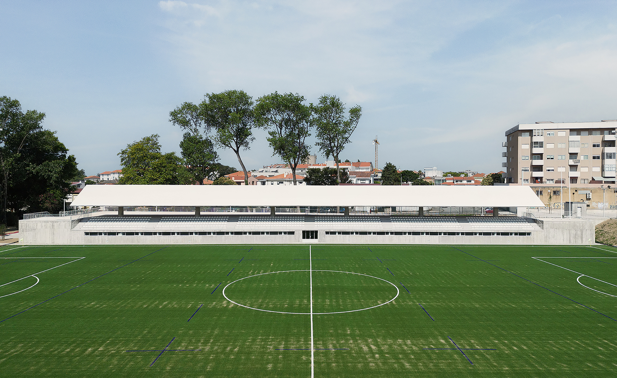 Concrete grandstand structure with horizontal window bands overlooking a football pitch with mature trees beyond