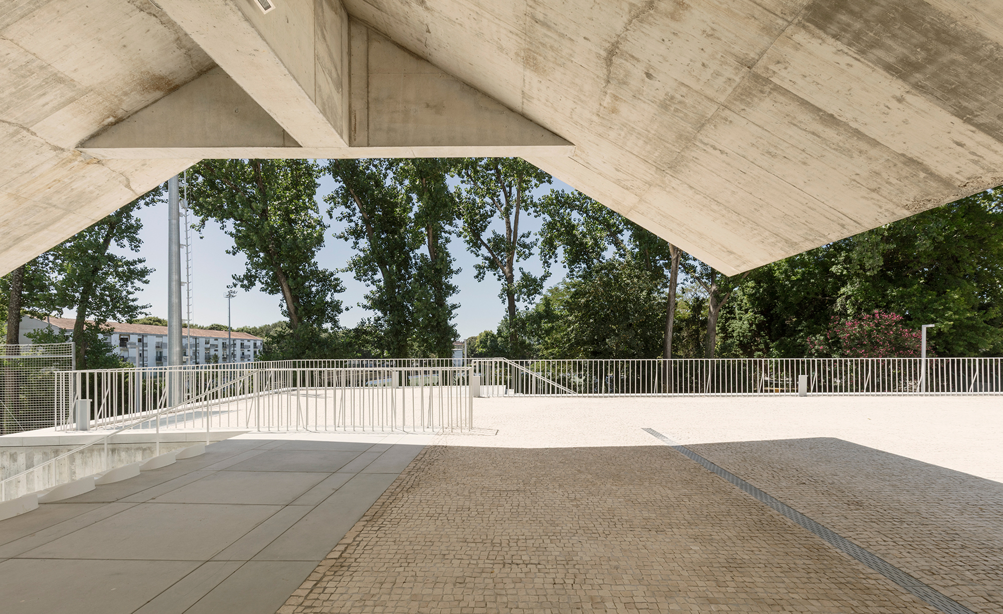 View from beneath the angular concrete canopy framing trees and perimeter fencing in bright daylight
