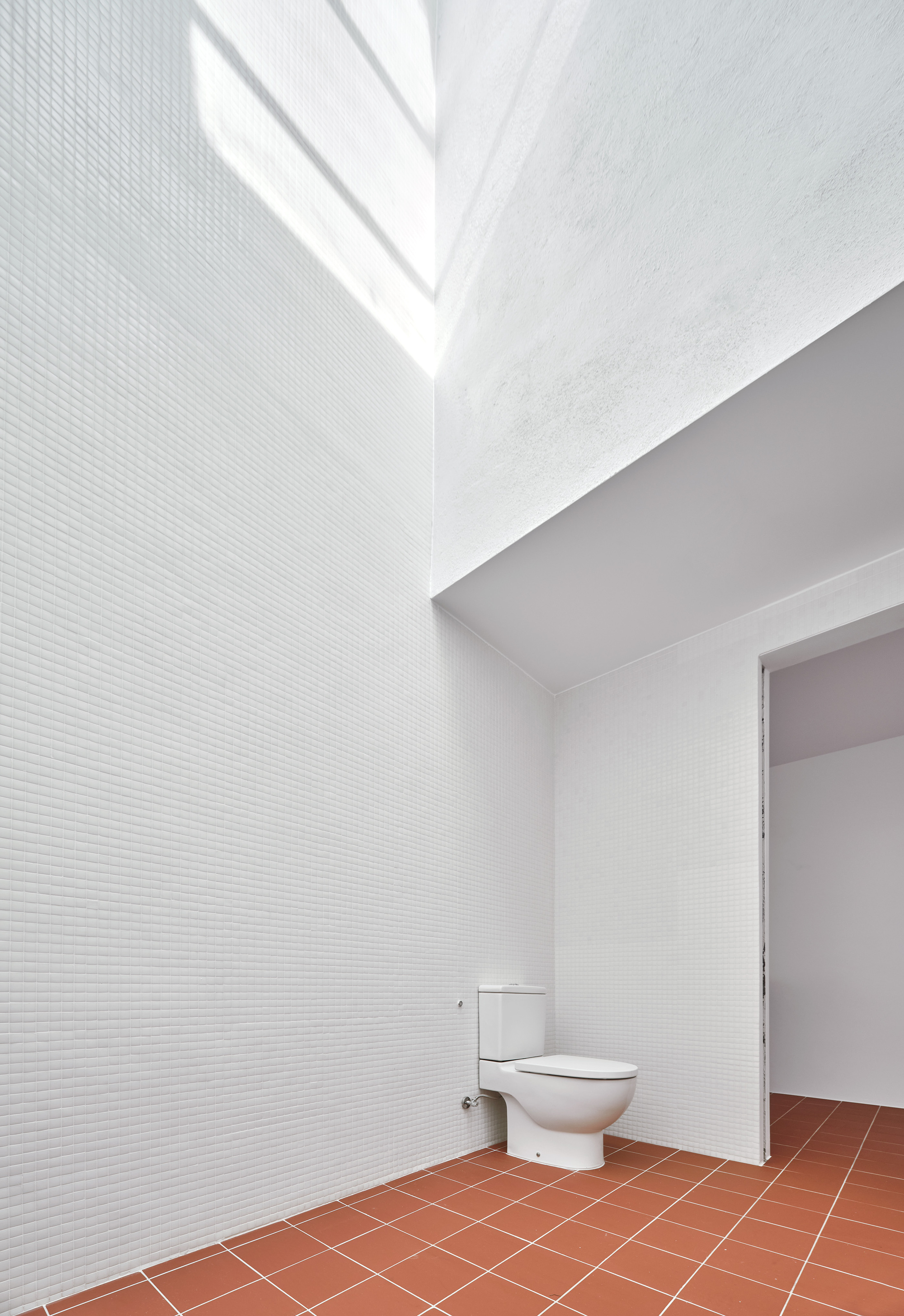 White-tiled bathroom with clerestory window casting angular shadows above terracotta floor and toilet fixture