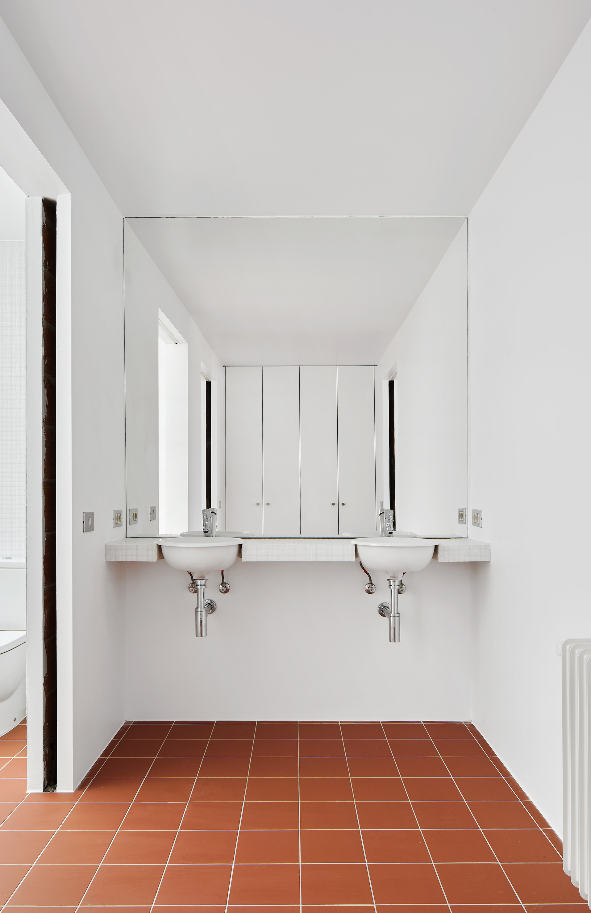 White bathroom with terracotta tile floor, wall-mounted twin sinks and large mirror reflecting cabinet doors