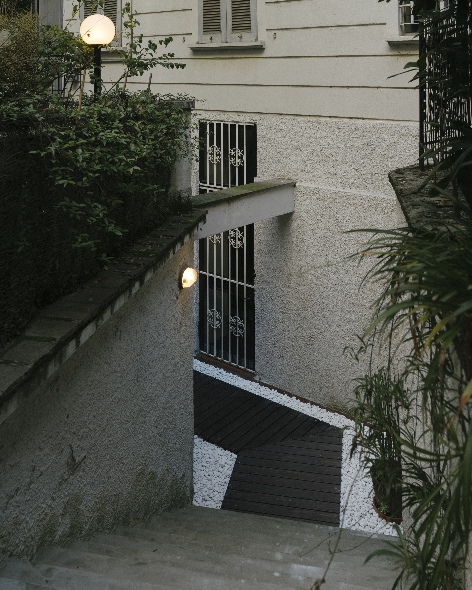 Top-down view of sunken courtyard with timber decking, white gravel, metal grilles, and wall-mounted spherical lights