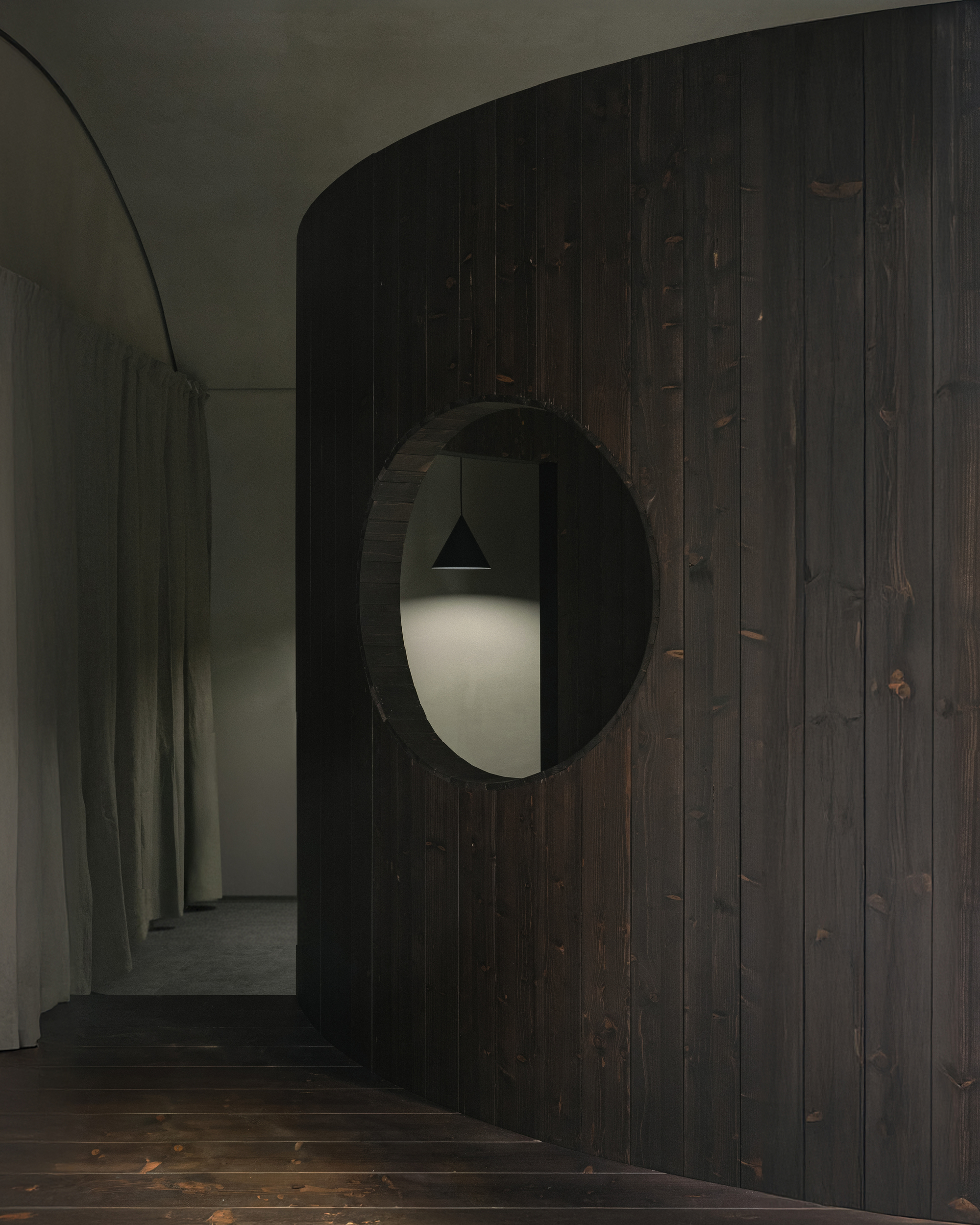 Curved dark timber wall with circular portal opening framing a pendant light and doorway beyond