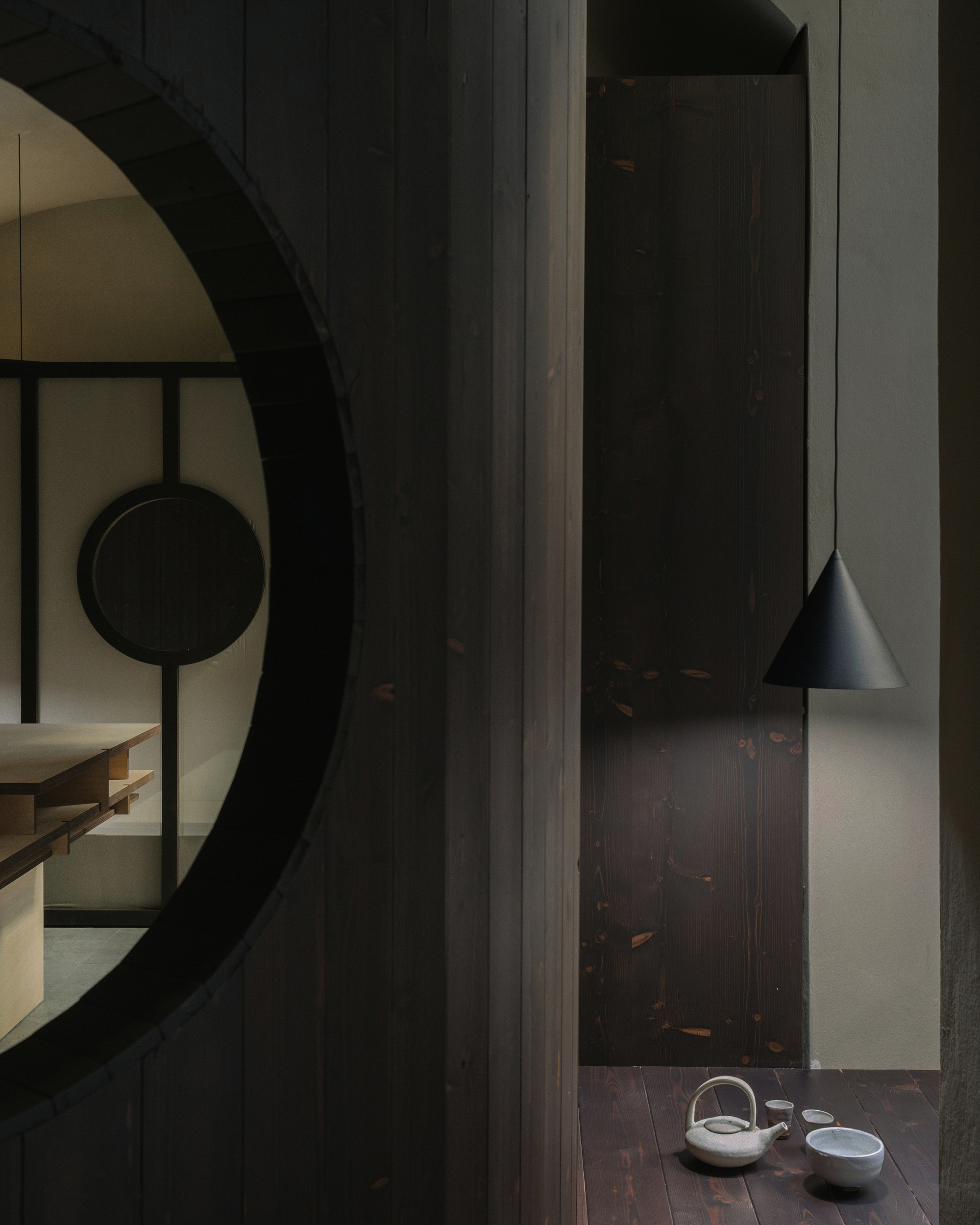 Dark timber wall with circular portal opening and conical pendant light above ceramic tea set
