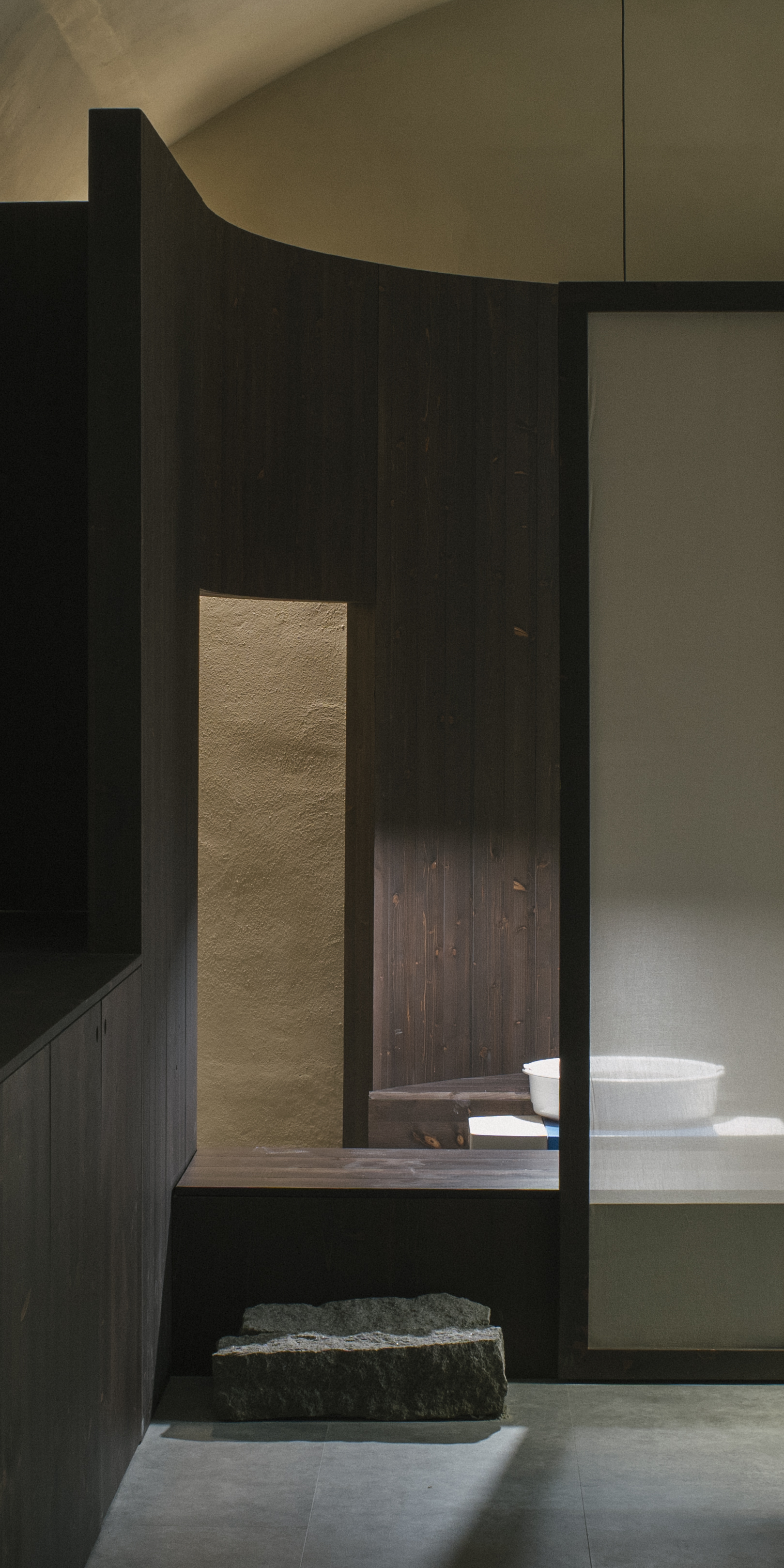 Layered timber partitions with narrow aperture revealing stone basin and shelving beyond