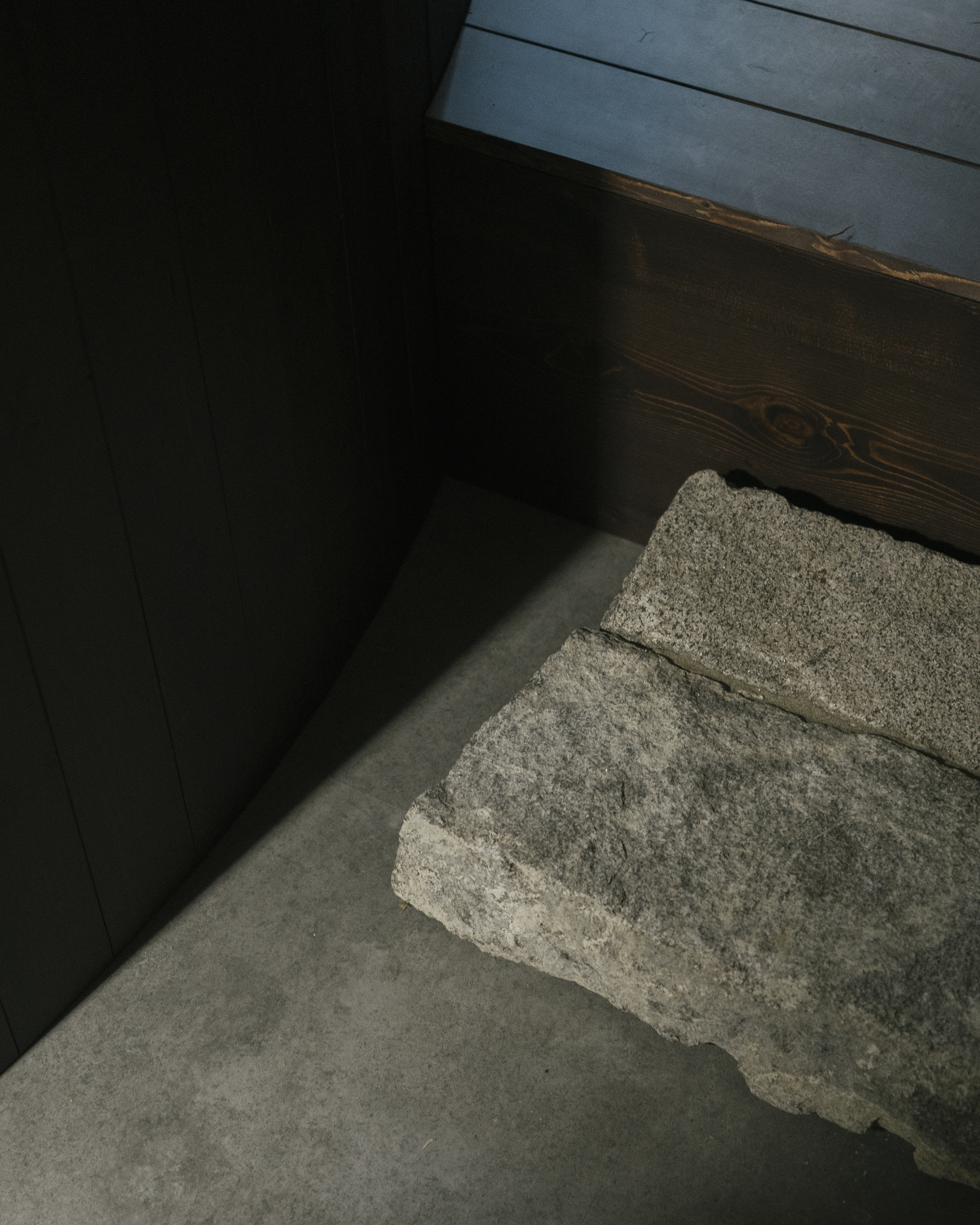 Grey concrete floor meeting a rough stone block and dark timber wall in soft daylight