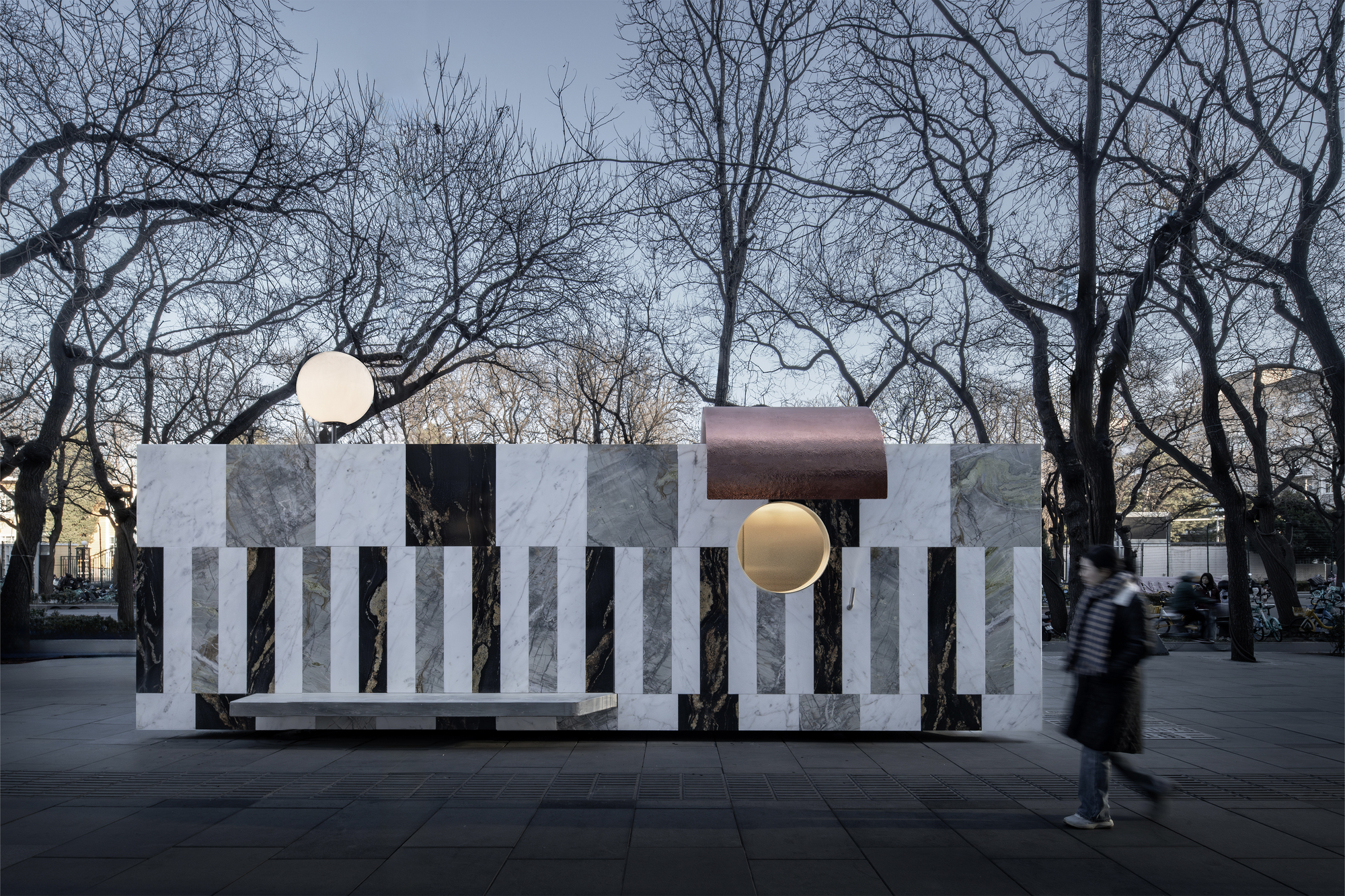 Freestanding pavilion with alternating marble panels and copper canopy in a public plaza with bare winter trees