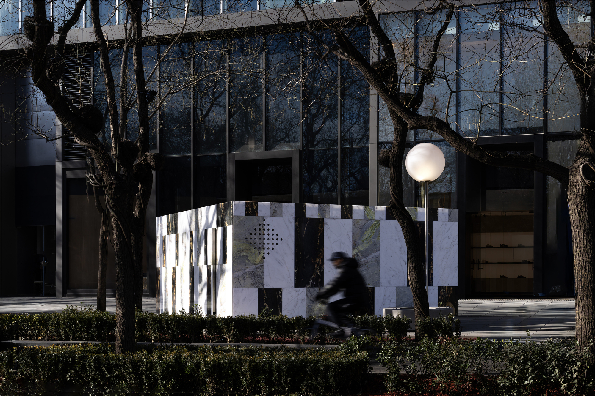 Striped marble screen wall with circular brass portal and white globe light as a cyclist passes by