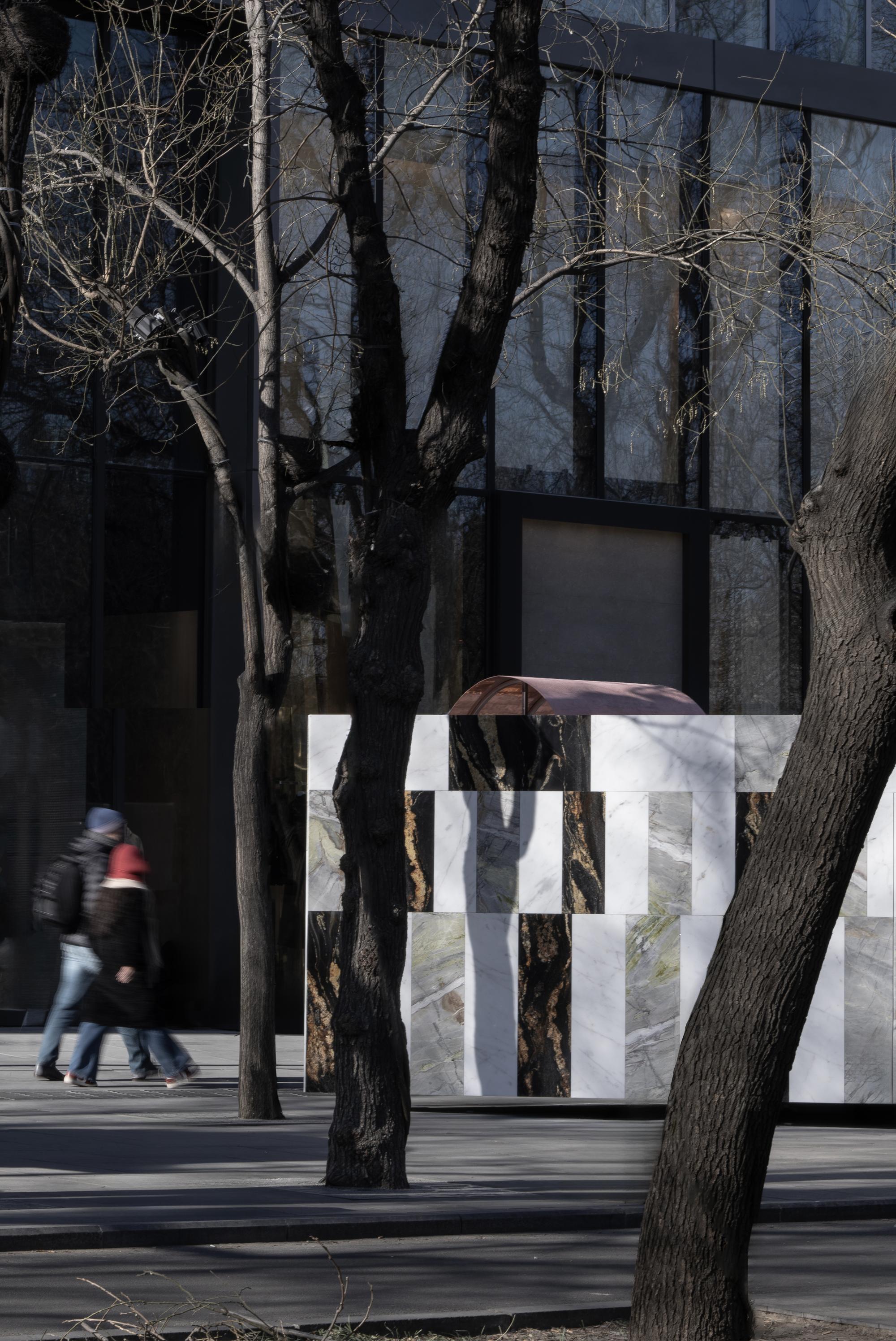 Striped stone wall installation with copper arch element as pedestrians pass beneath bare tree shadows