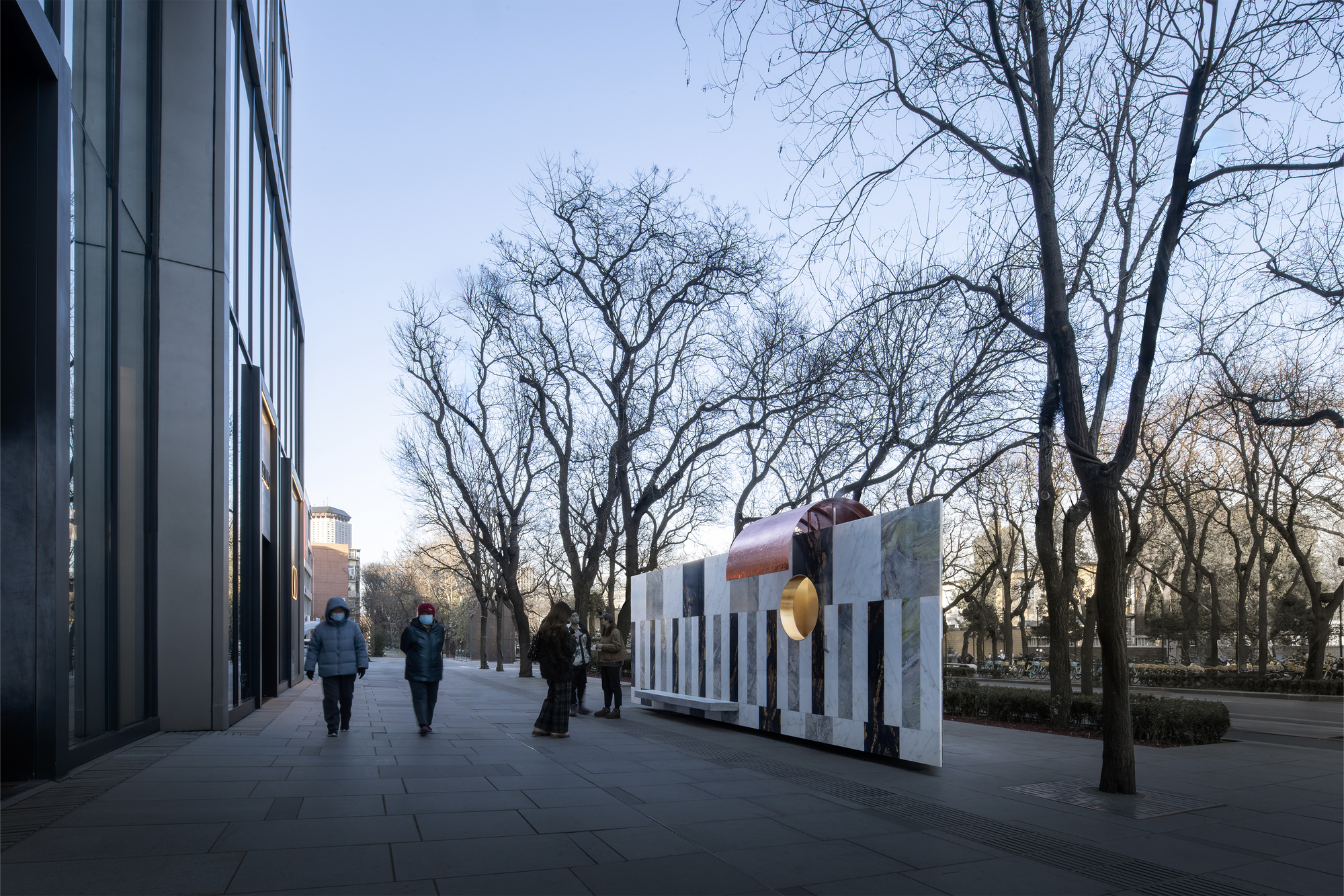 Pedestrians walking along paved plaza with freestanding stone wall and copper tube sculpture at sunset
