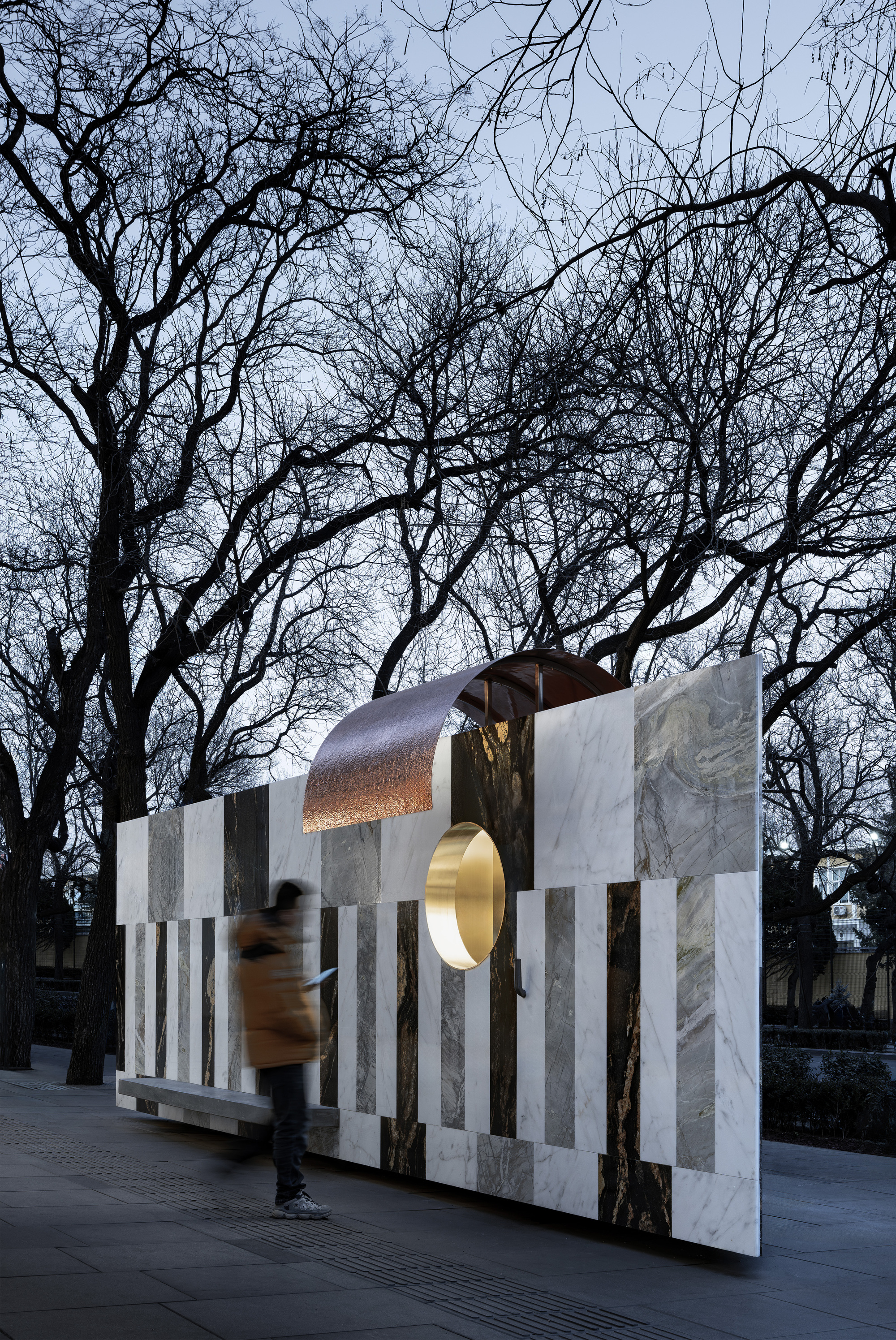 Freestanding wall of vertical stone stripes with copper canopy and pedestrian passing through at twilight