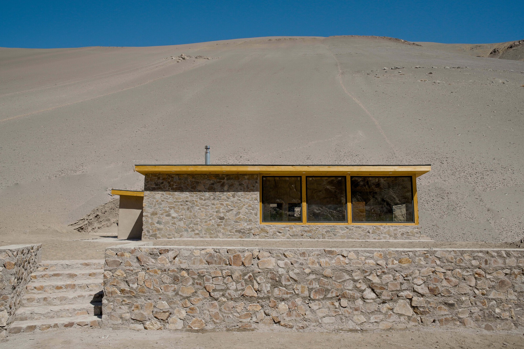 Stone wall structure with yellow-framed ribbon windows set against a barren desert hillside under bright blue sky