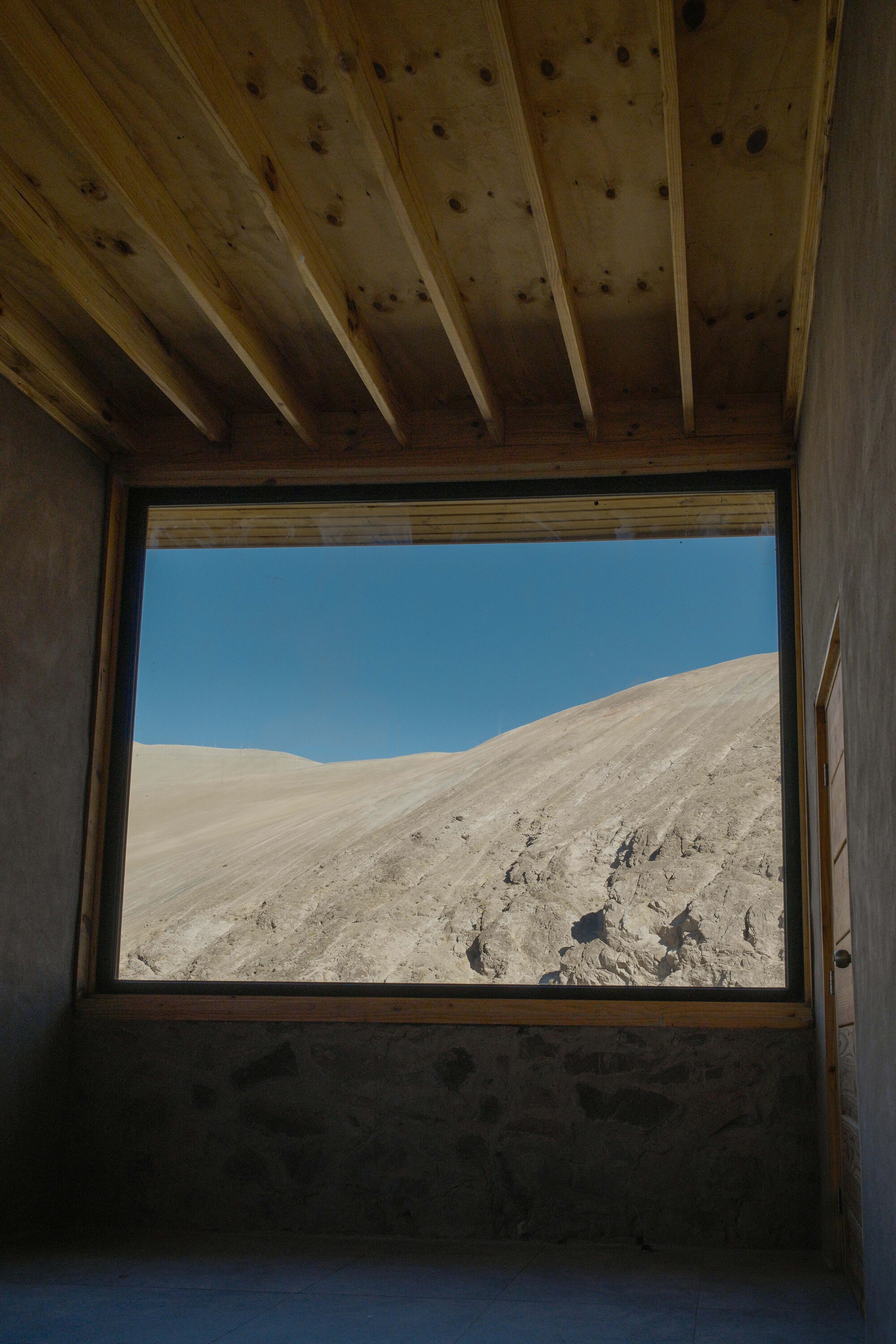 Square window framing the desert hillside beyond with exposed timber joists overhead