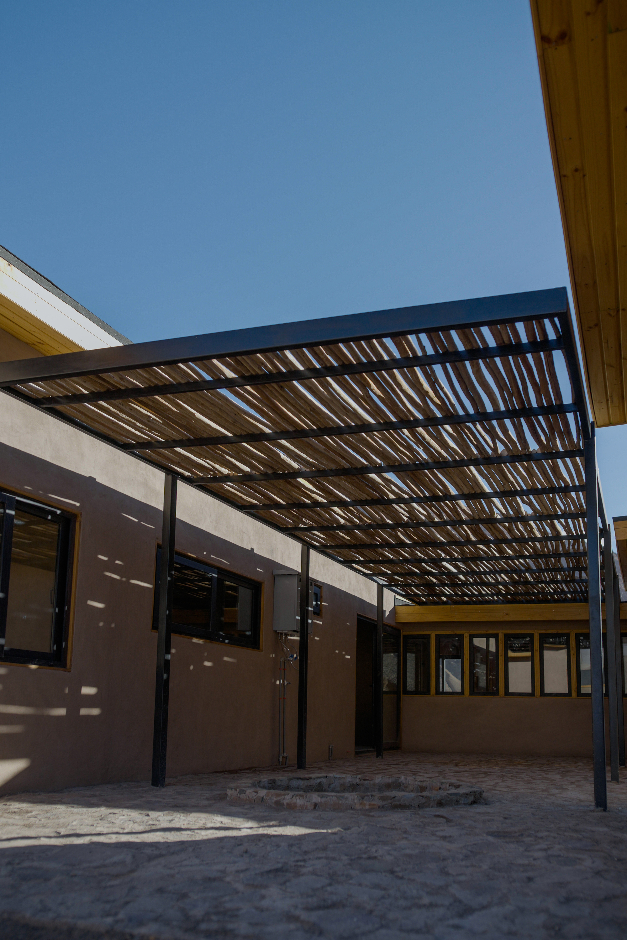 Sheltered courtyard with slatted steel overhead canopy filtering daylight onto plastered walls and stone paving