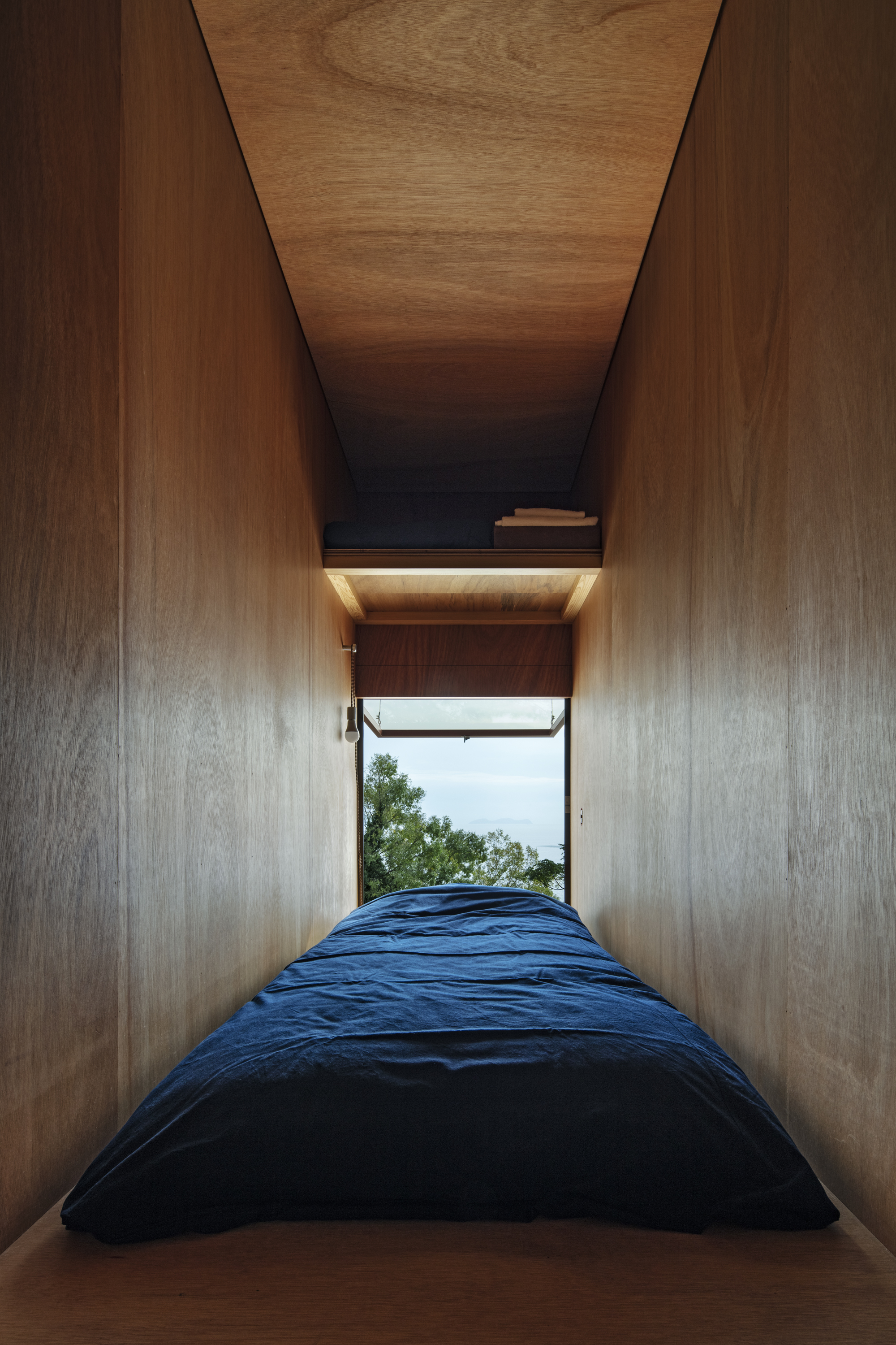 Narrow sleeping nook with plywood walls and ceiling opening to a window with ocean view