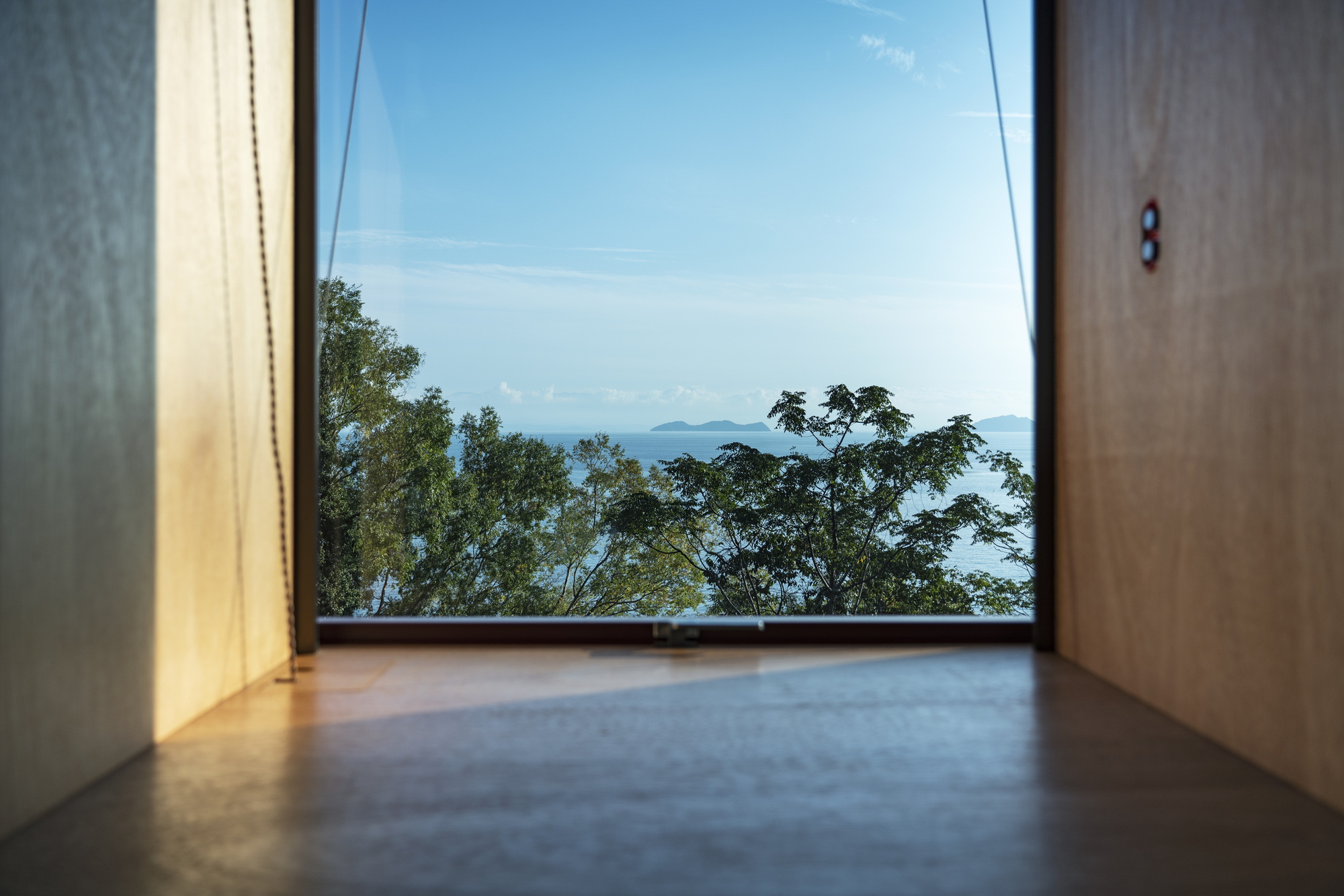 Floor-level view through a window framing treetops and ocean islands under clear sky