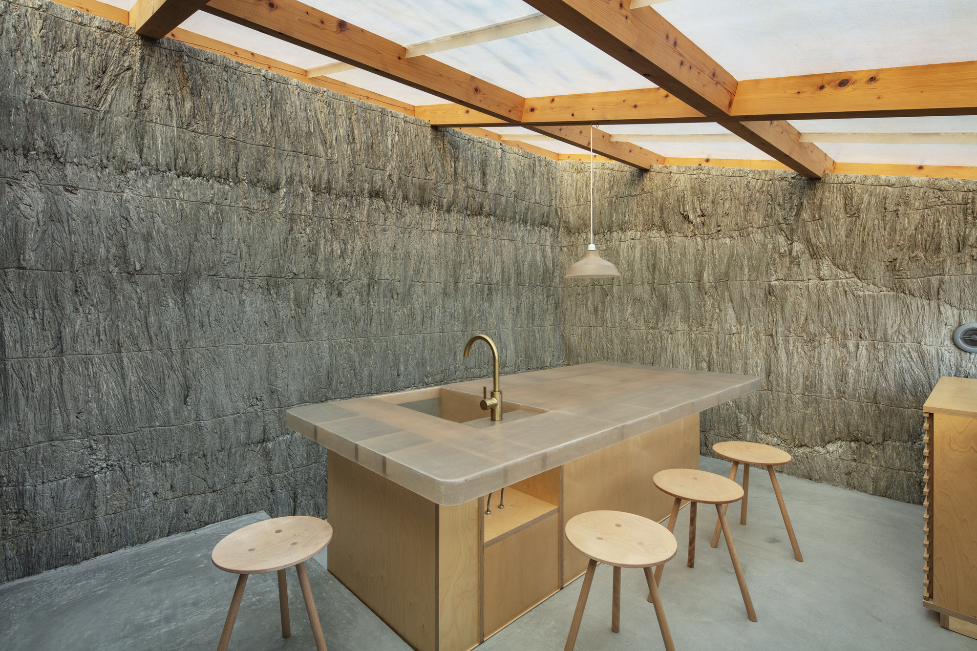 Kitchen island with wooden stools beneath exposed timber roof structure and textured concrete walls