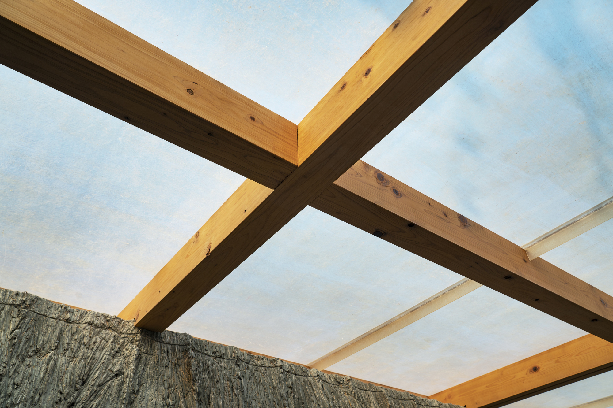 View upward through crossing timber beams framing the hempcrete ceiling against blue sky