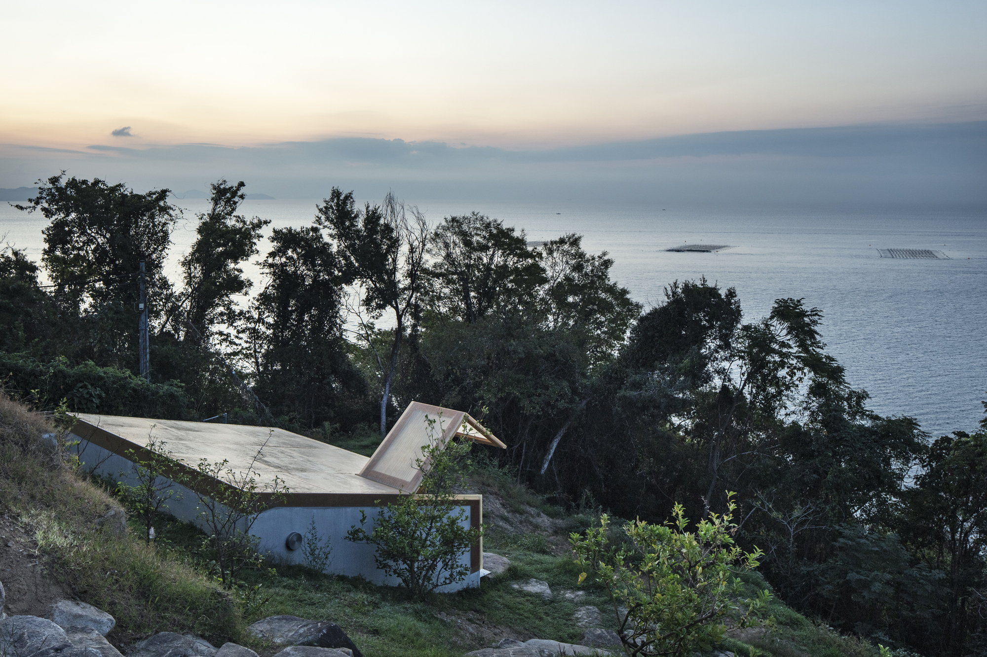 Folded concrete roof nestled into the hillside vegetation above the ocean at dusk
