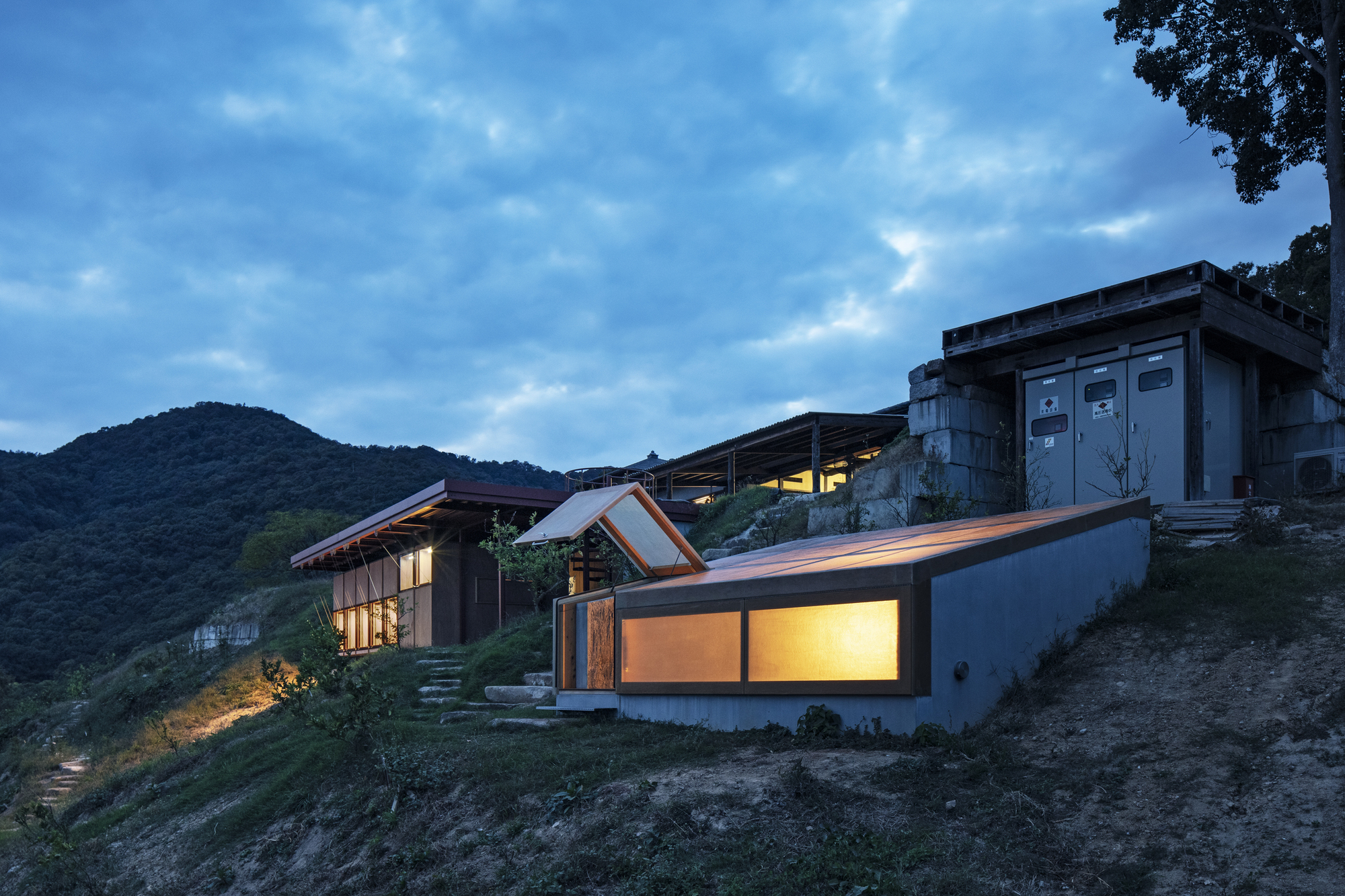Cluster of volumes with angled roofs and illuminated glazing on the sloped site at twilight