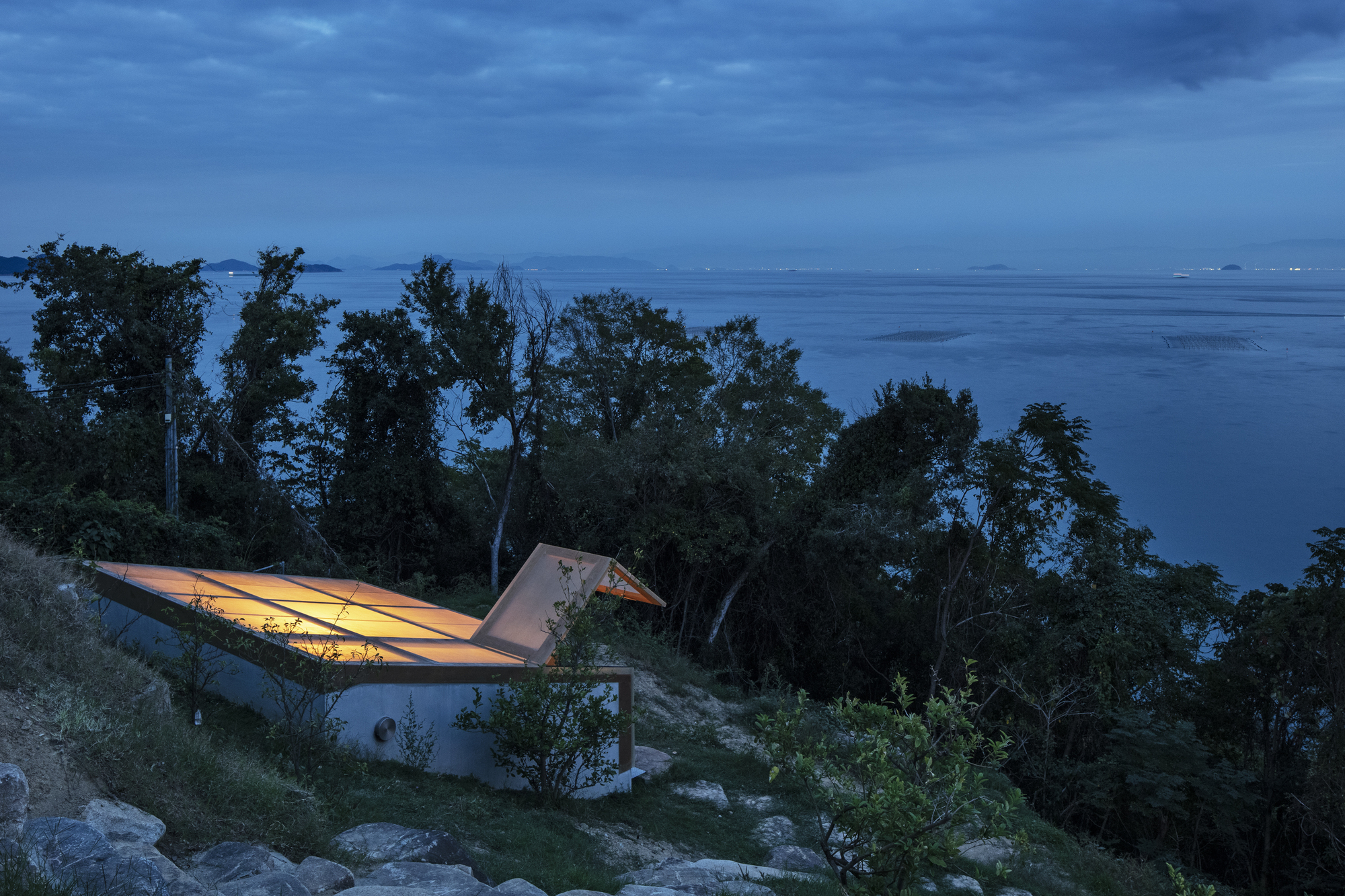 Cantilevered roof illuminated from within overlooking the forested coastal hillside at night