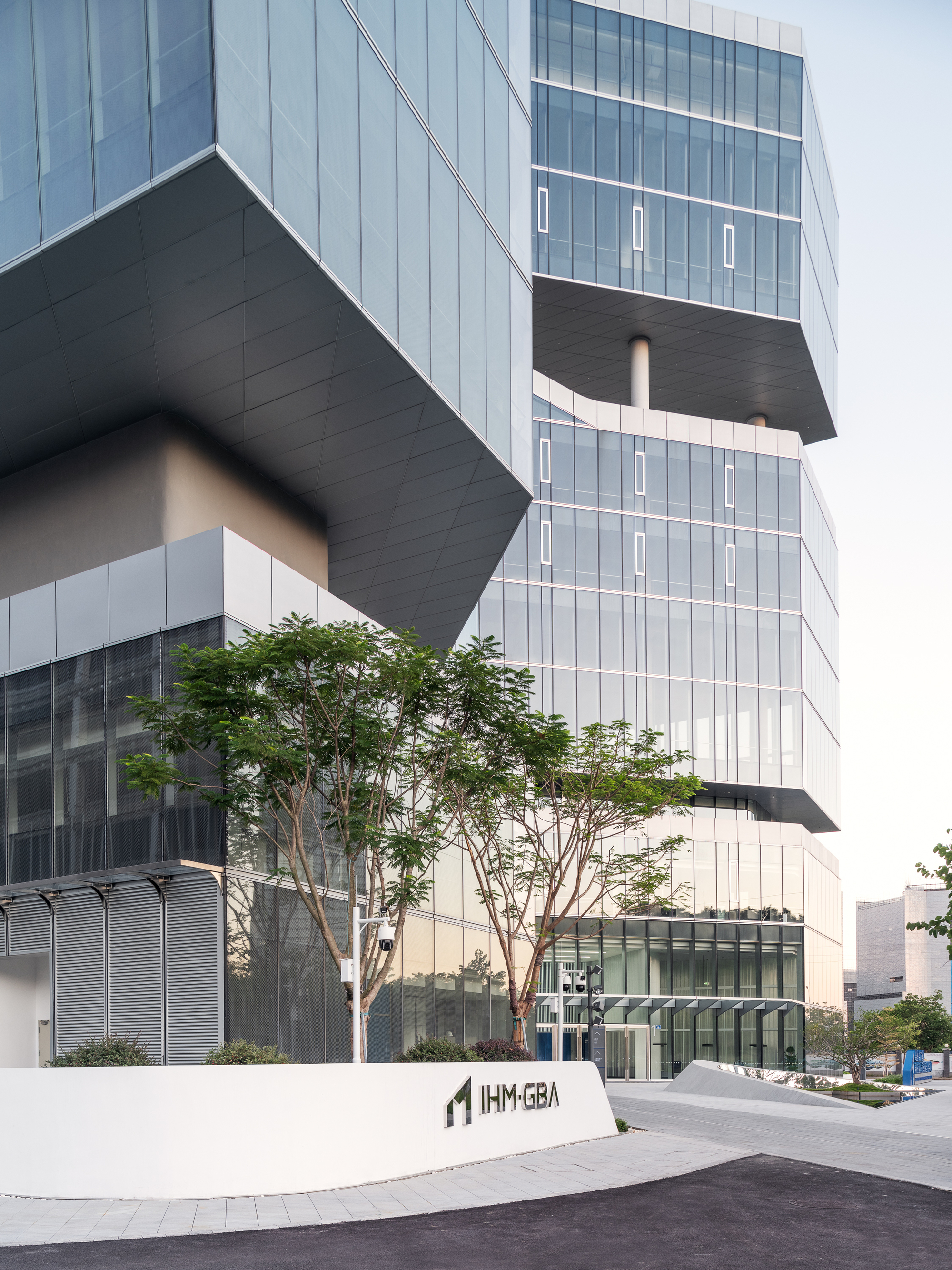 Street view of the stacked glass volumes with cantilevered dark slabs and a mature tree at the entrance