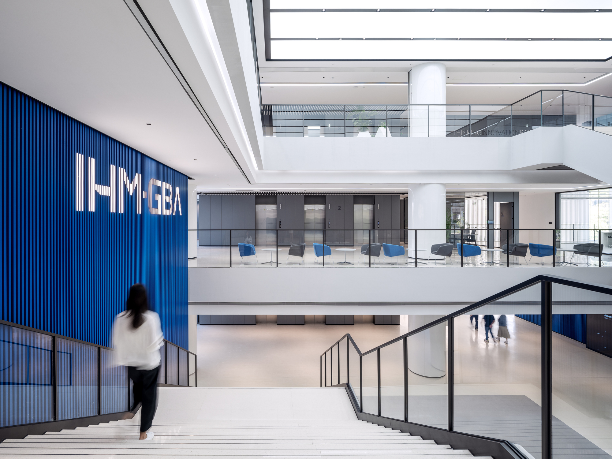 Multi-story atrium with white stairs descending past a blue corrugated wall and a person walking