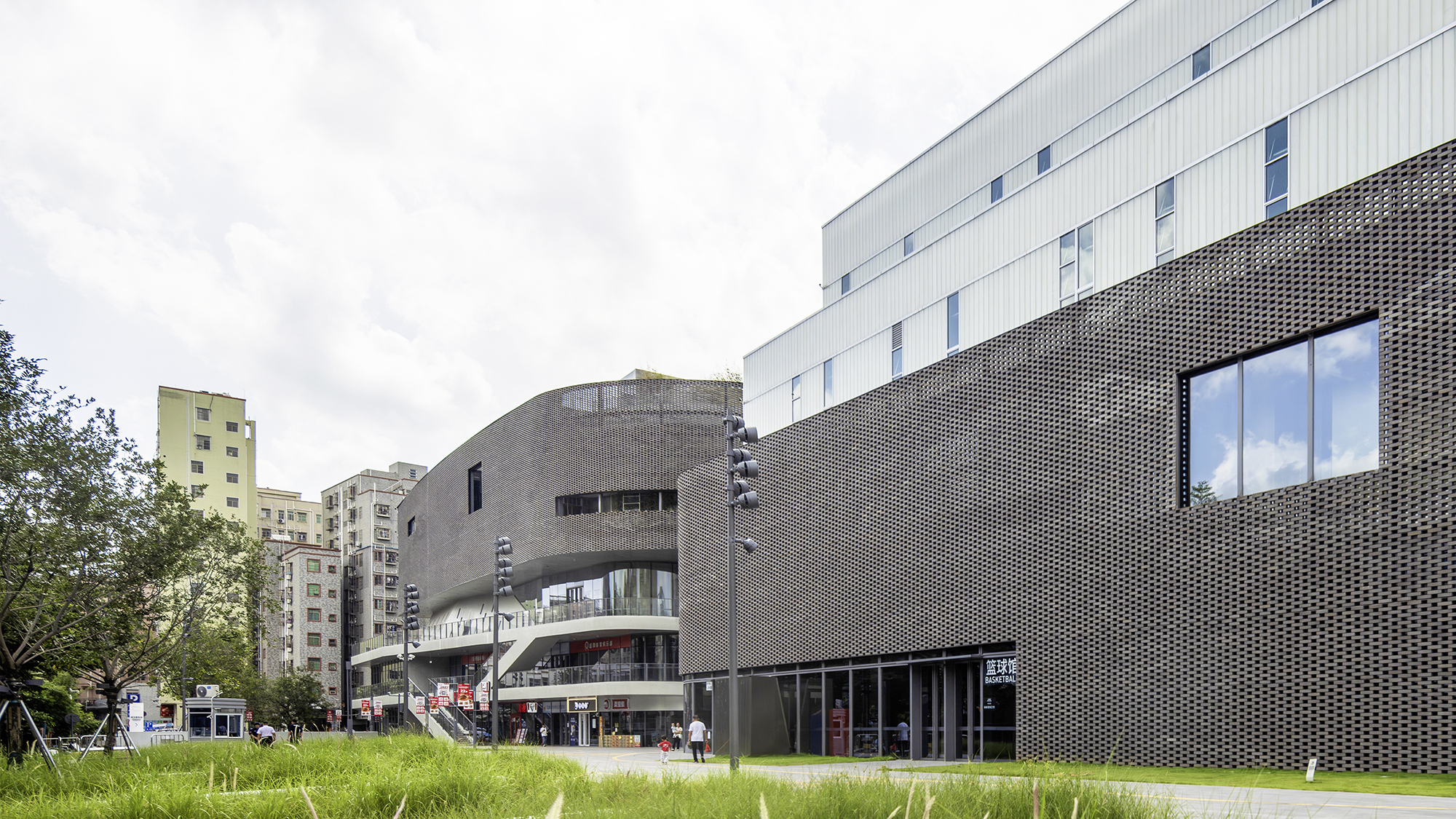 Perforated metal facade wrapping curved volumes beside a grassy lawn with trees on an overcast day