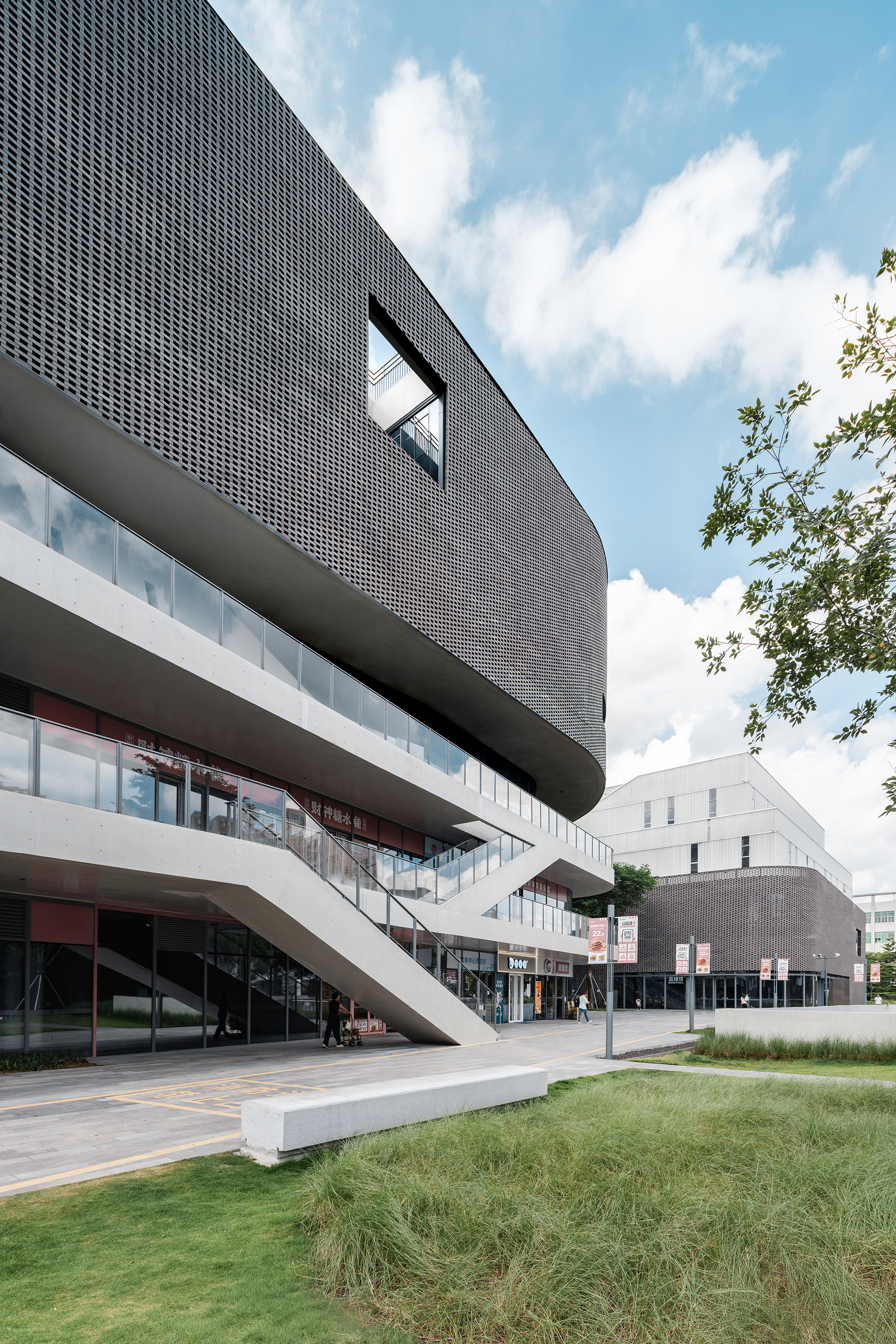Cantilevered concrete bands extending over glazed entry beneath a dark perforated metal screen with blue sky