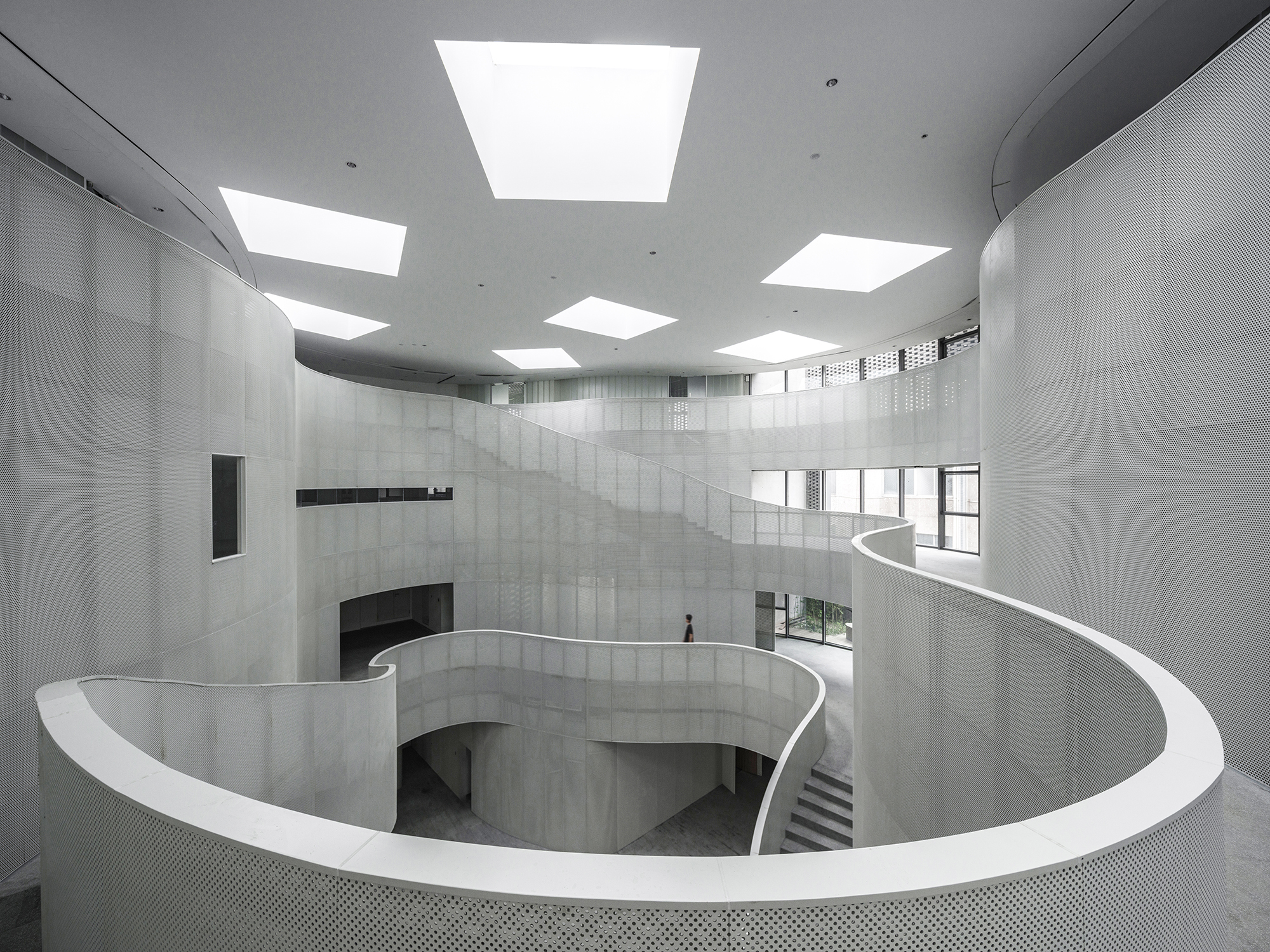 Spiraling white balustrade and concrete walls beneath square skylights in a multi-story atrium with a lone figure